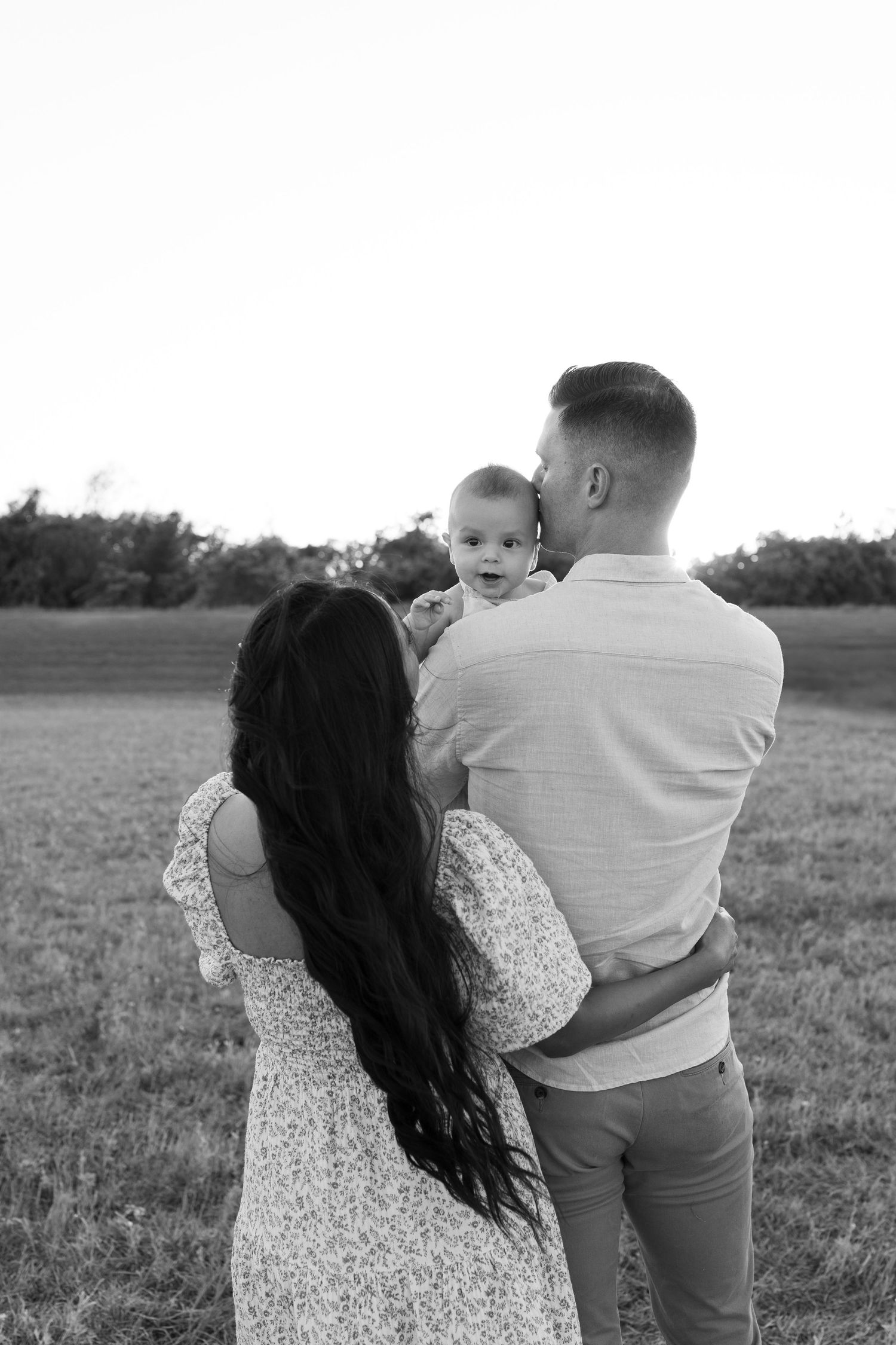 Black and white photo of a couple in a field lovingly holding their baby while gazing at each other outdoors.