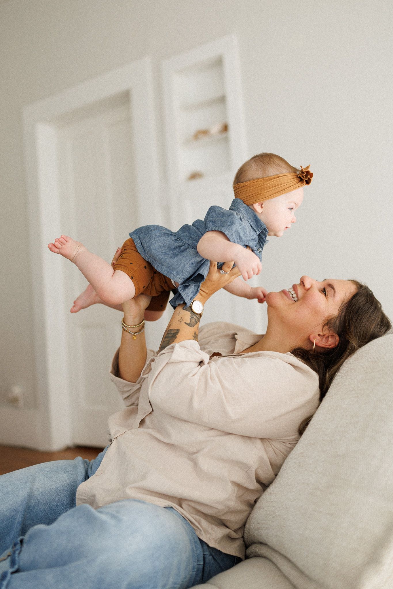 A happy mother lifts her smiling baby girl while relaxing on a couch in a bright living room.