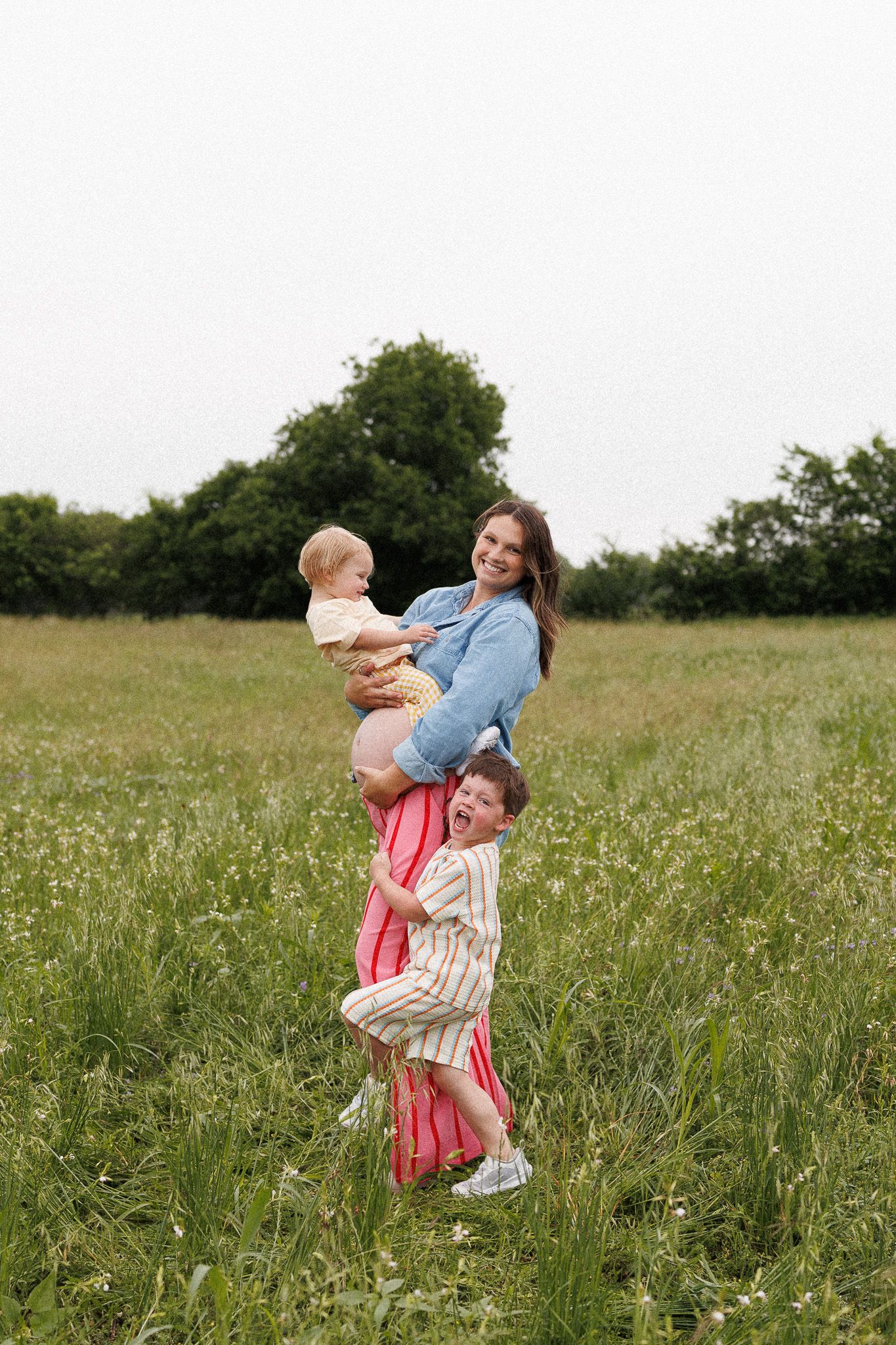 Pregnant woman holding baby while toddler hugs her legs in a green meadow field outdoors.