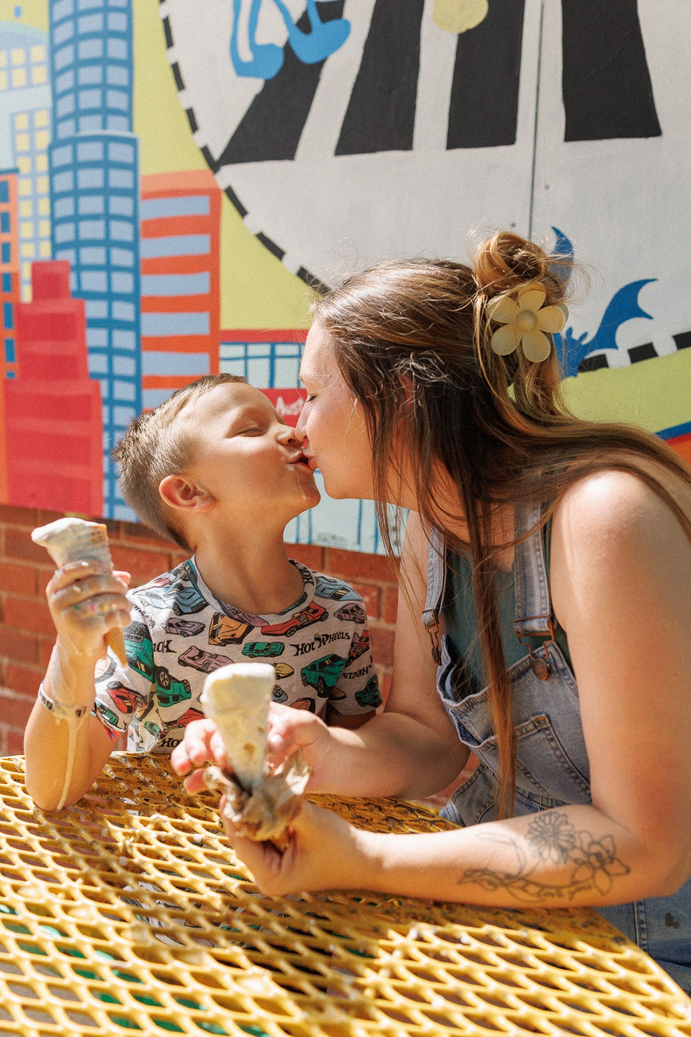 A mother and child share a kiss while enjoying ice cream cones at an outdoor table with colorful mural backdrop.