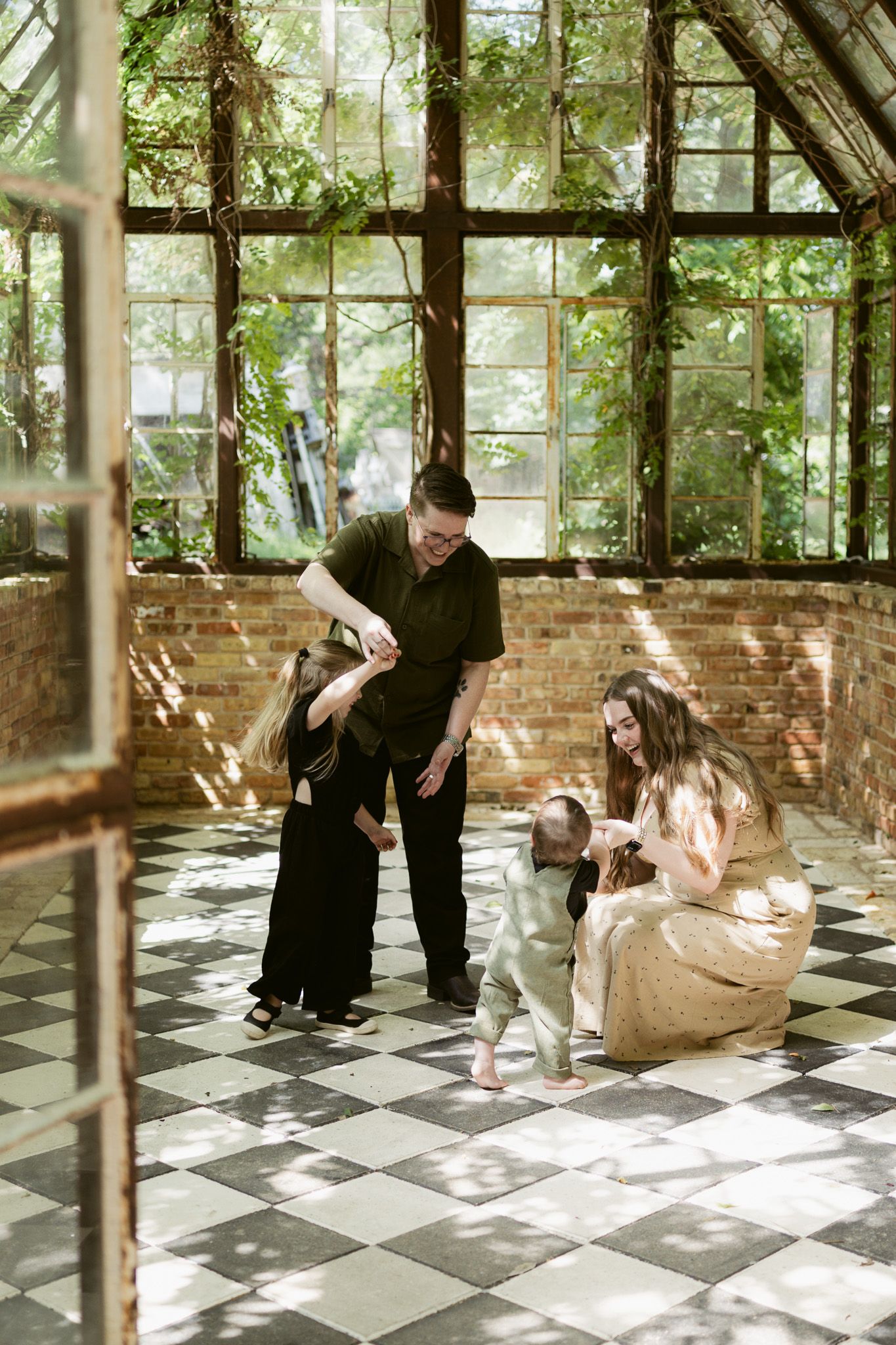 Family playing together on checkered floor in sunlit greenhouse with large windows and lush greenery.