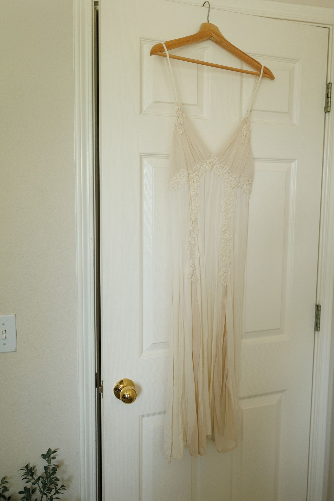 A delicate cream-colored slip dress with spaghetti straps hangs from a wooden hanger on a white door.