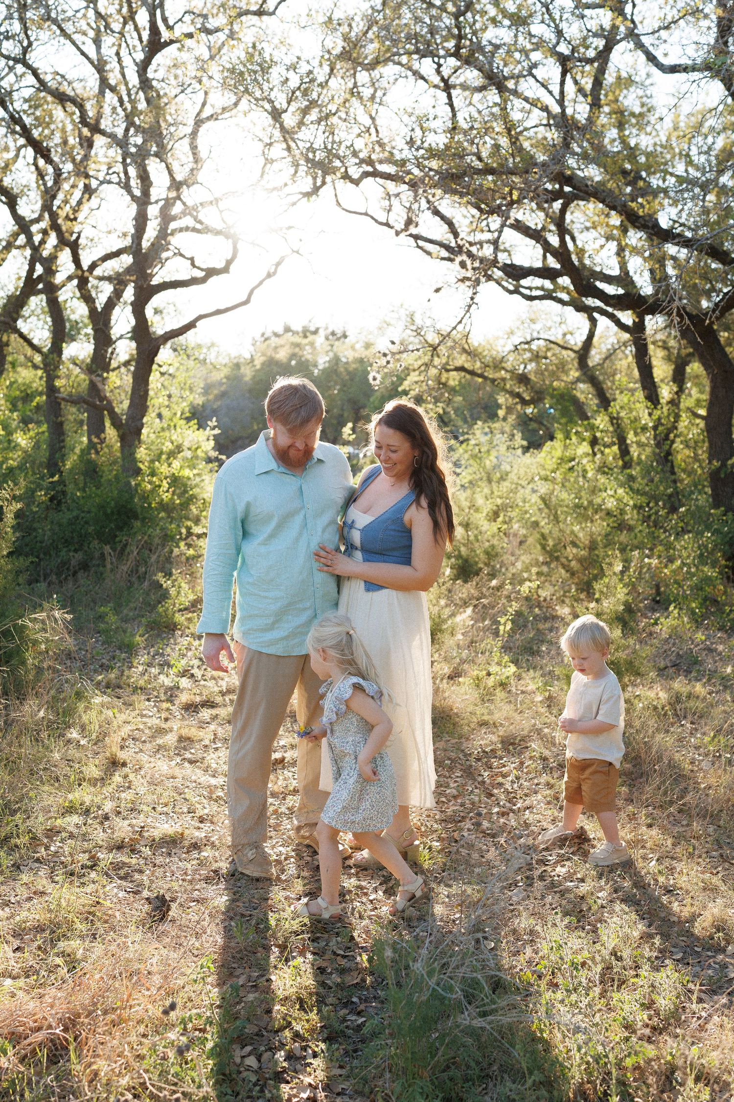 A family of four walks along a sunlit woodland path, with a pregnant mother, bearded father, and two young children.