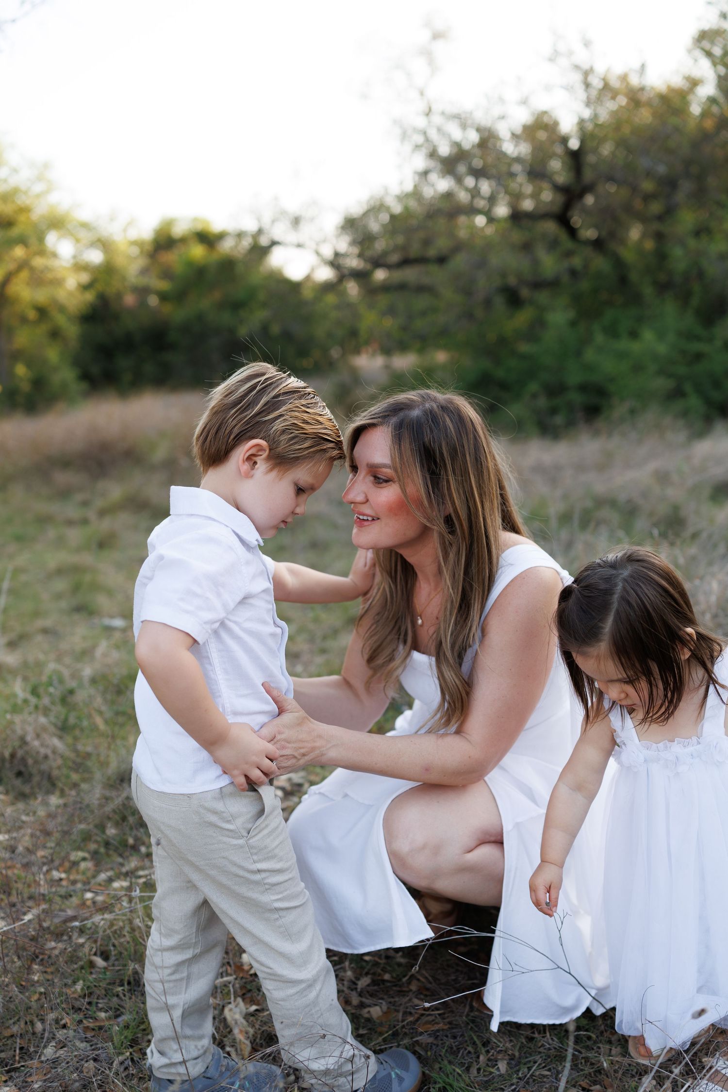 A mother in a white dress crouches outdoors with her young son and daughter in a serene natural setting.