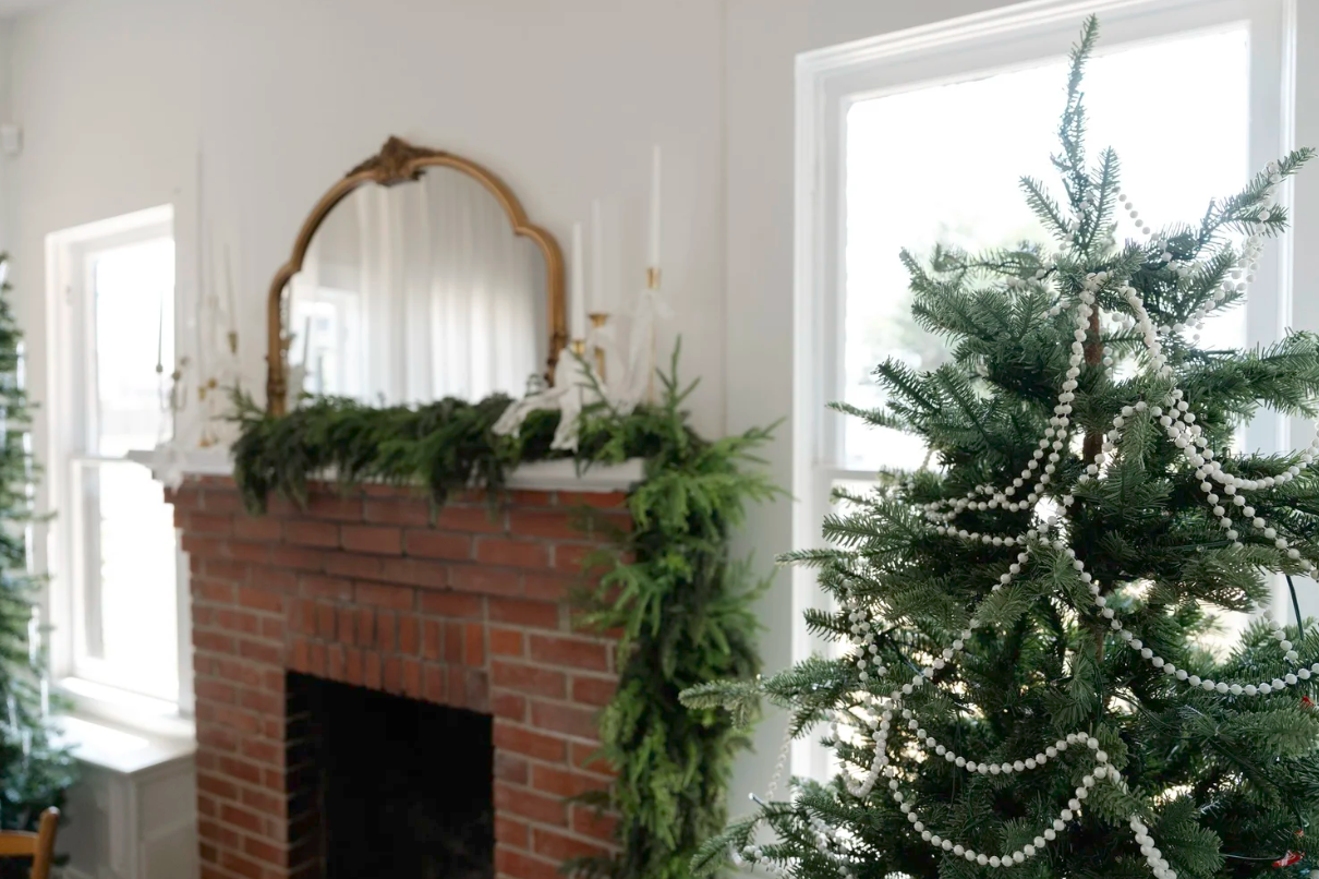 Christmas living room with brick fireplace, garland, gold mirror, and pearl-decorated evergreen tree near white windows.