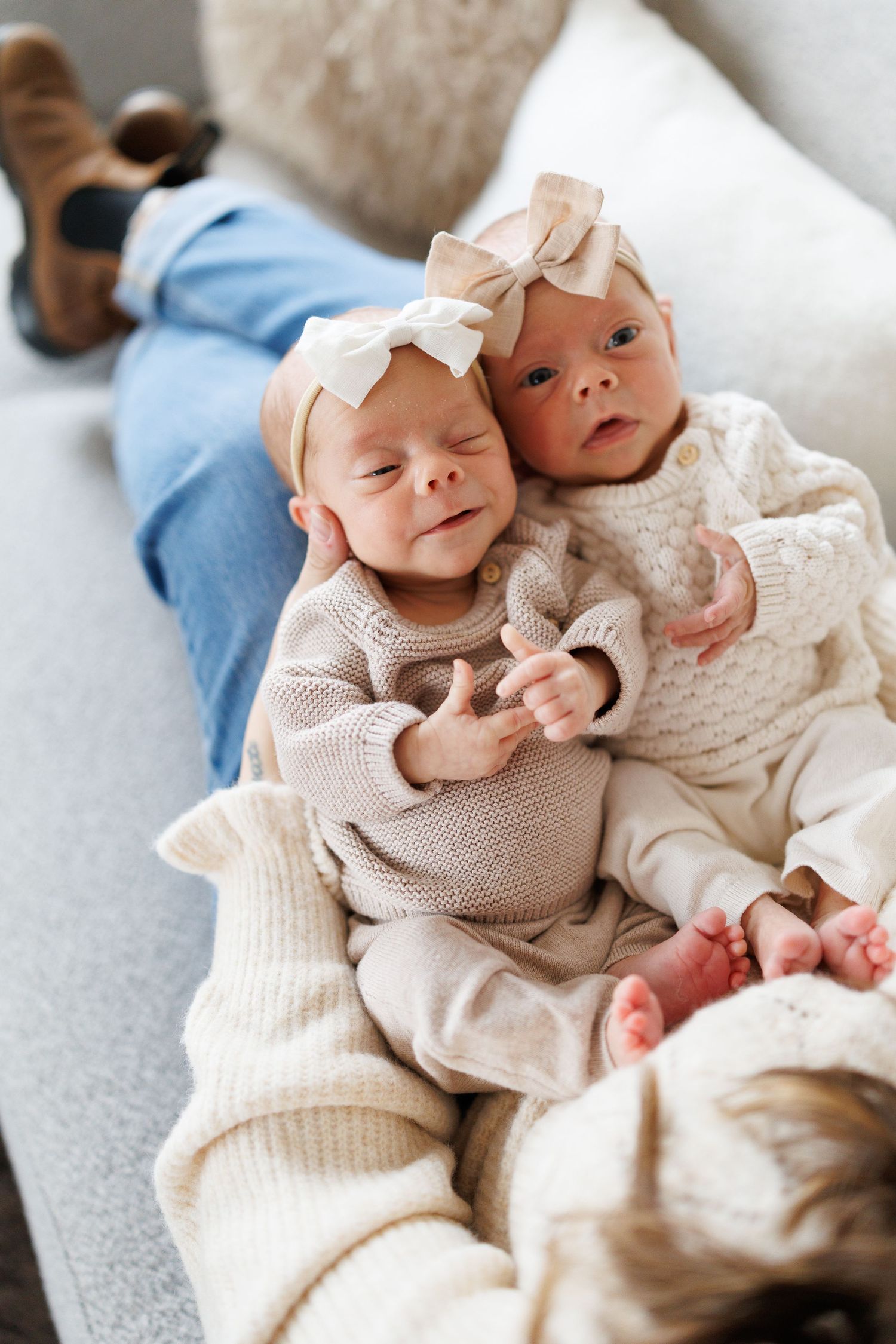 Twin babies in cream colored knit sweaters lie together on white bedding while smiling and cuddling.