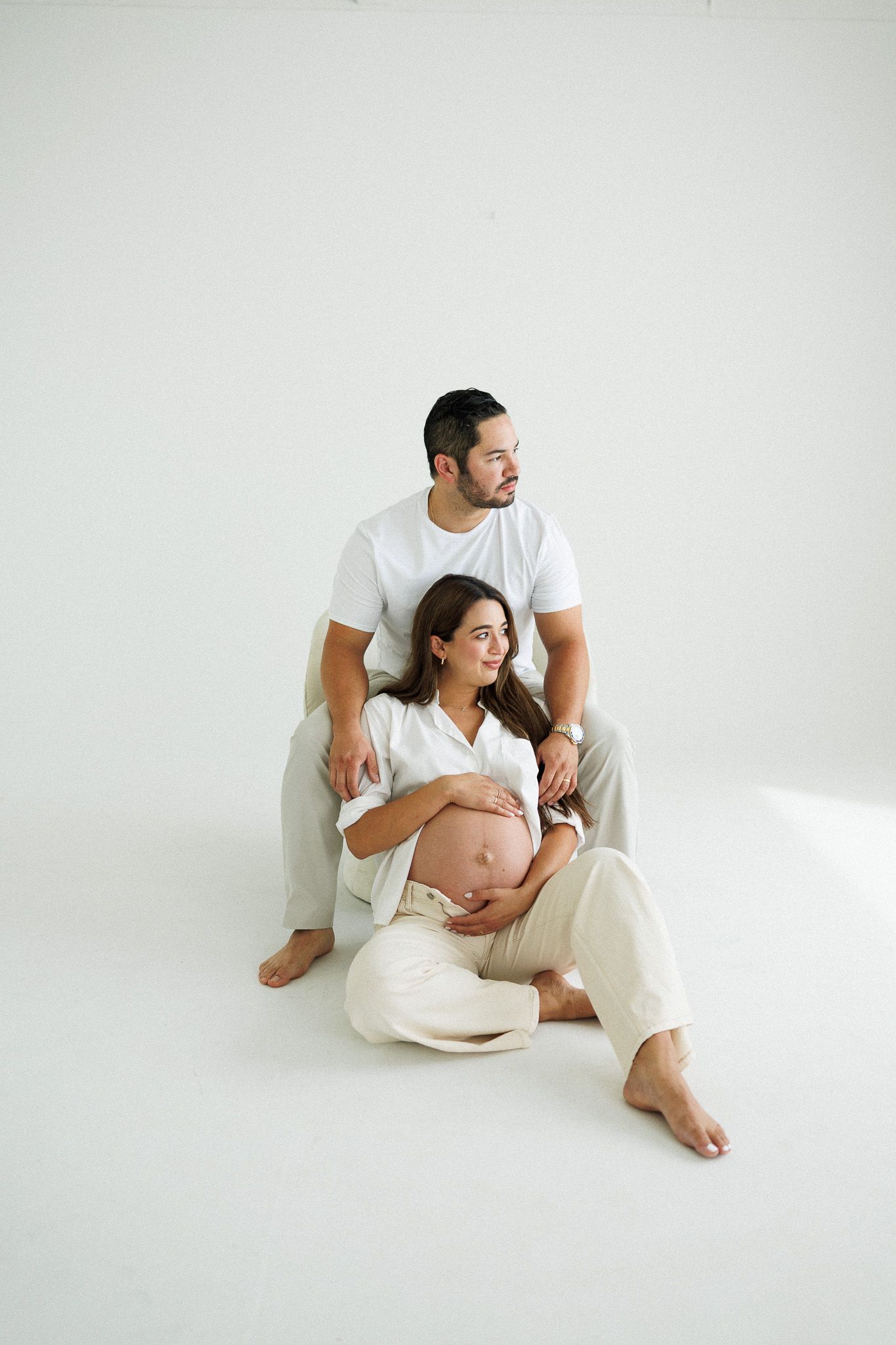 Pregnant couple sitting on white floor, man behind woman cradling her bare baby bump in a serene maternity photo.