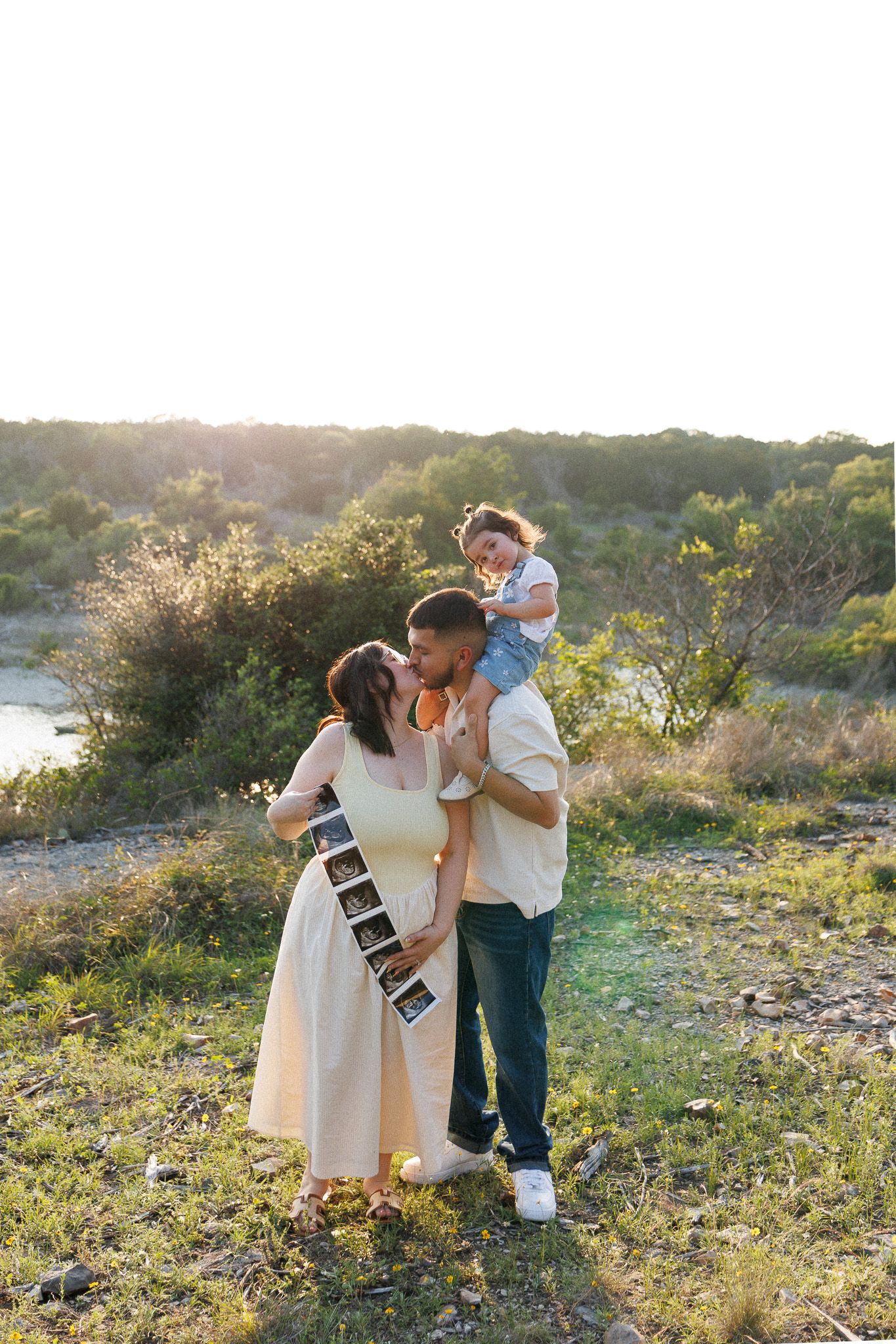 Pregnant woman holds ultrasound photos while man lifts toddler on shoulders outdoors at golden hour.