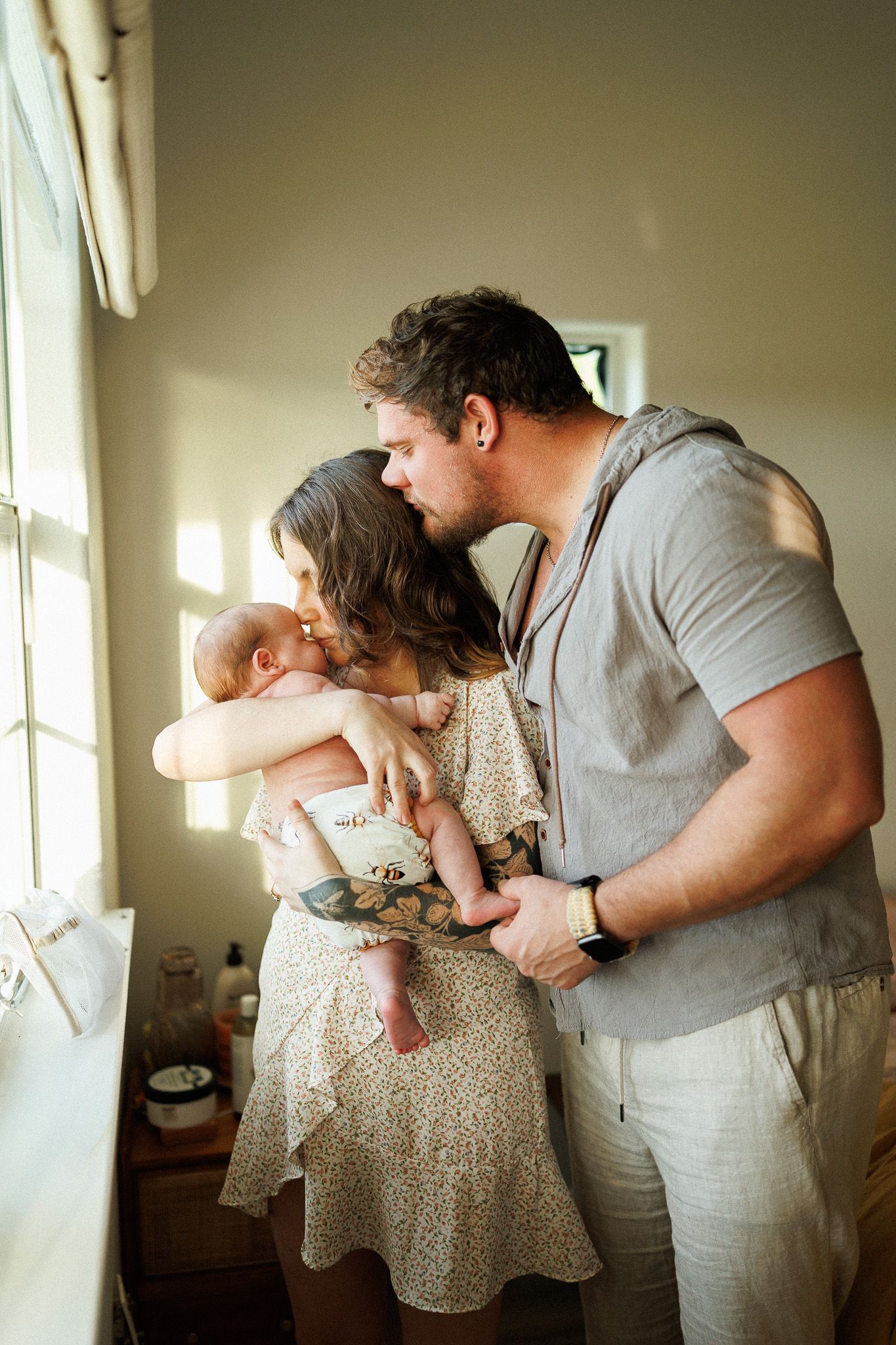 A couple shares a tender moment holding their newborn baby by a sunlit window indoors.