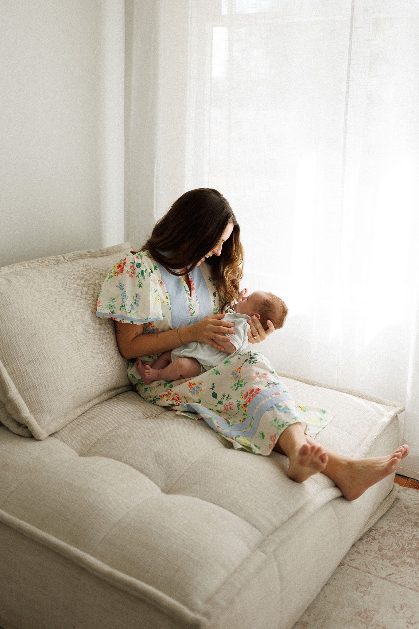 Mother in floral dress cradling newborn baby on a cream sofa by window.