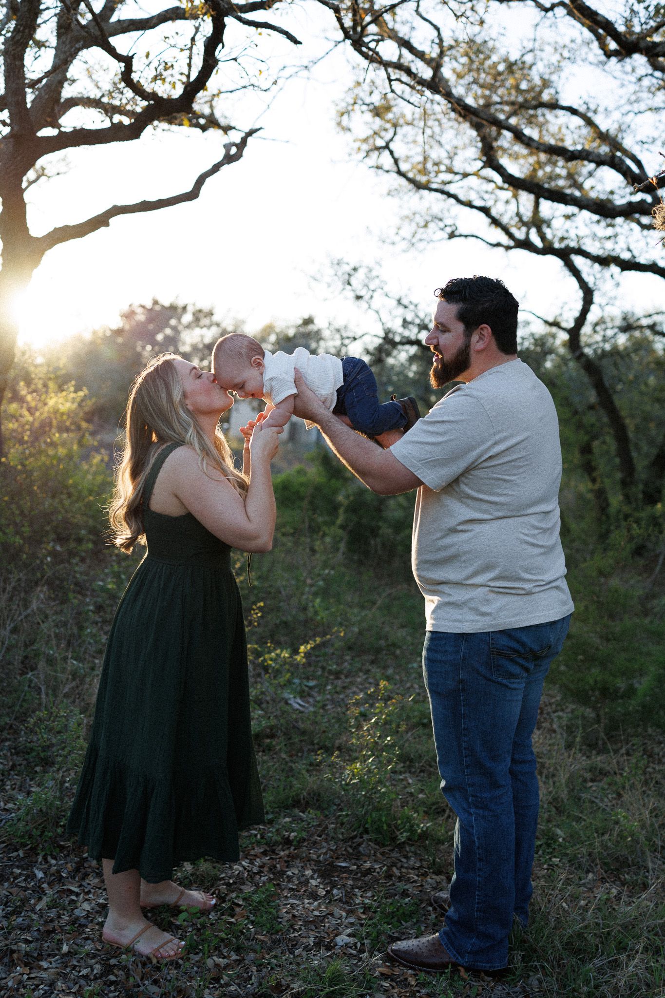 A couple shares a tender moment with their baby in a natural outdoor setting at sunset among oak trees.