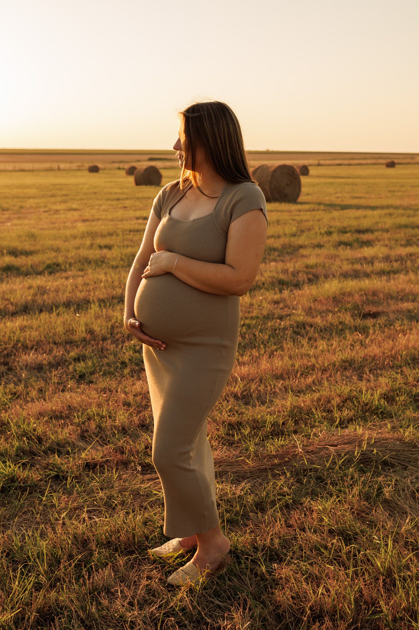 Pregnant woman in gray dress stands in golden field at sunset with round hay bales scattered in background.