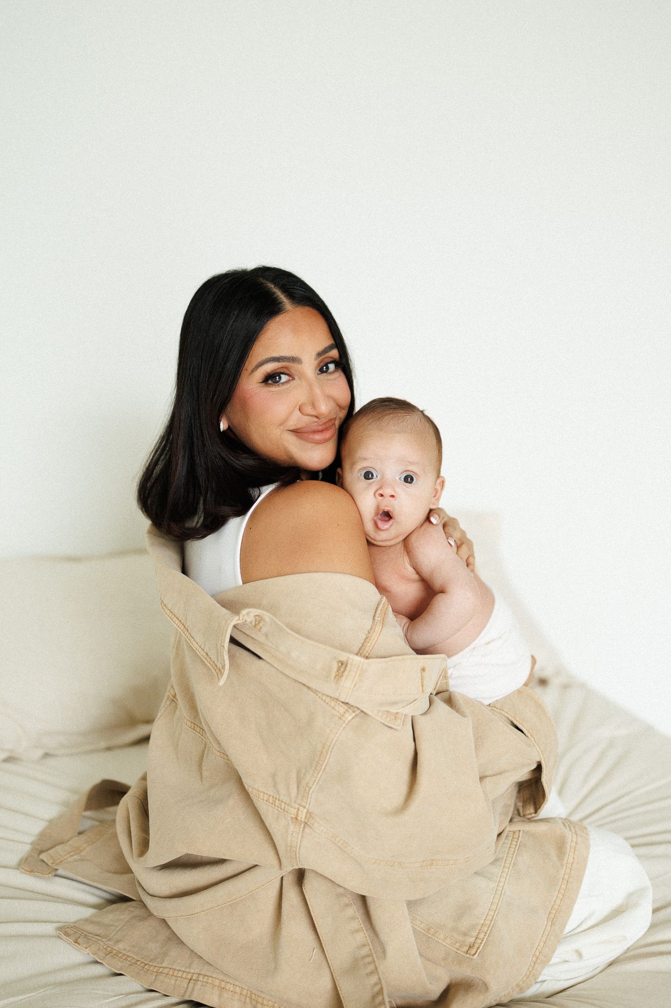 A parent and baby share an intimate moment while wrapped in a beige blanket against a white backdrop.