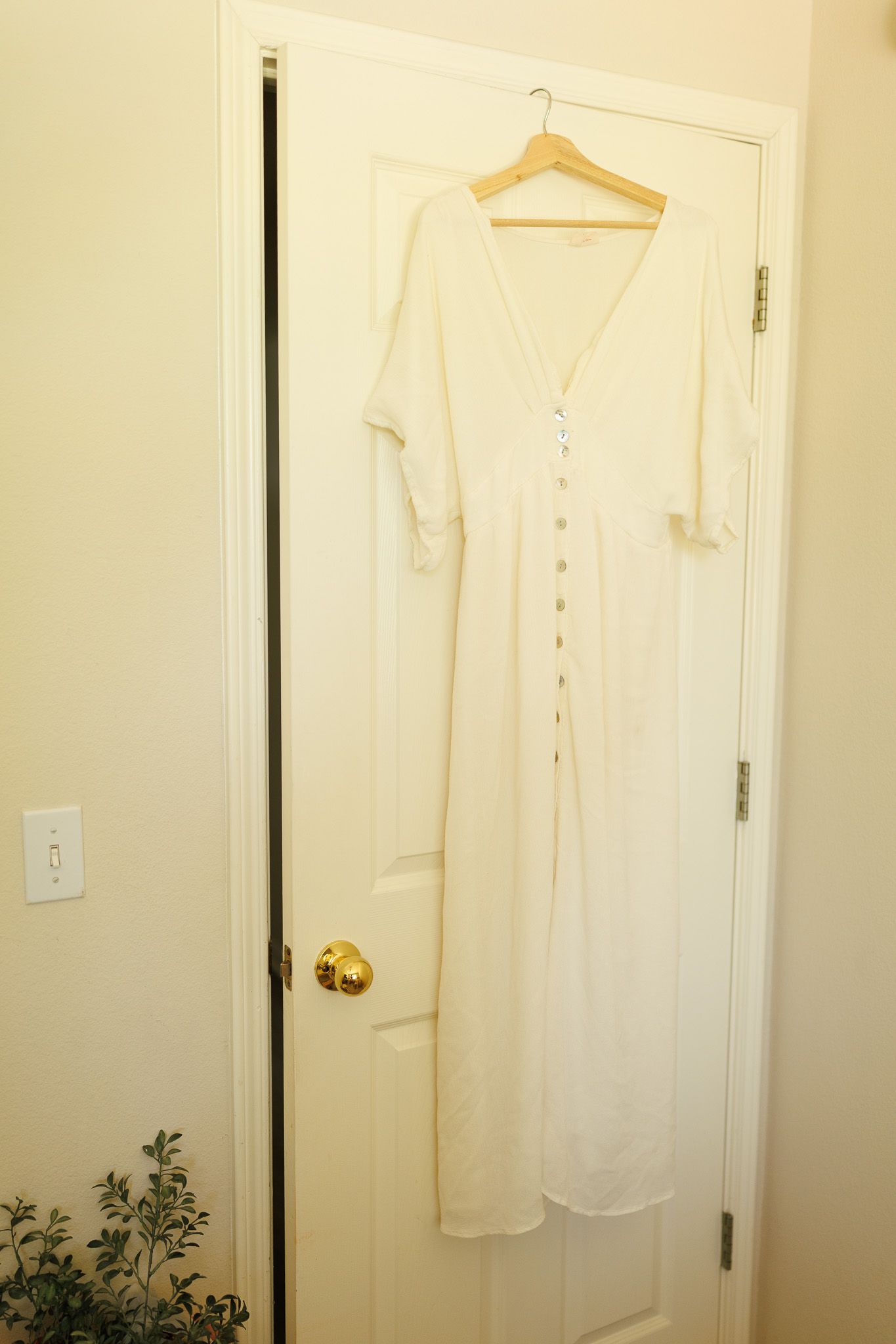 Long white button-up sleeveless dress shirt hanging on a wooden hanger against a cream-colored wall and door.