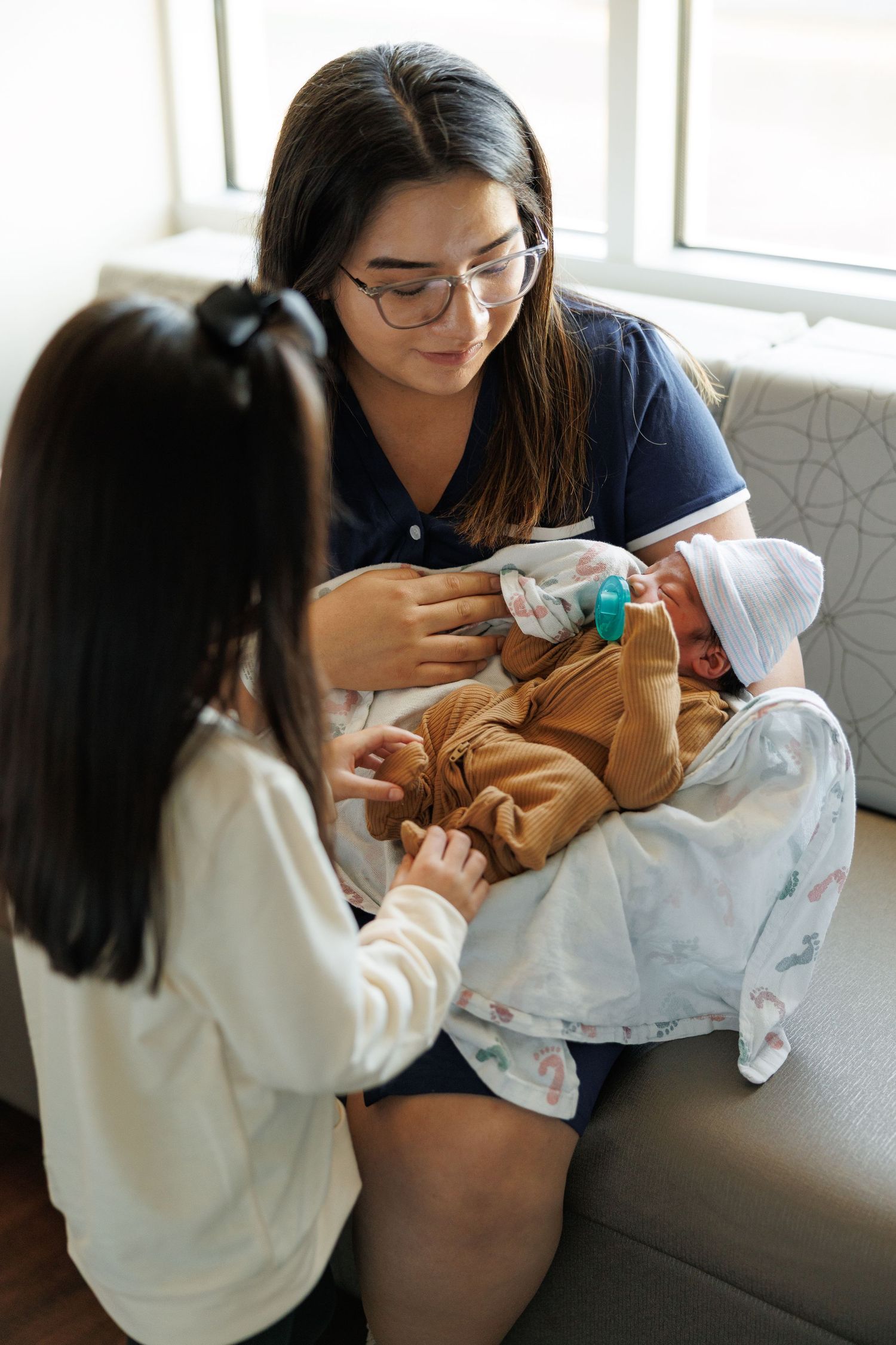 Two people sit on a gray couch and examine baby clothing or blankets together in a sunlit room.