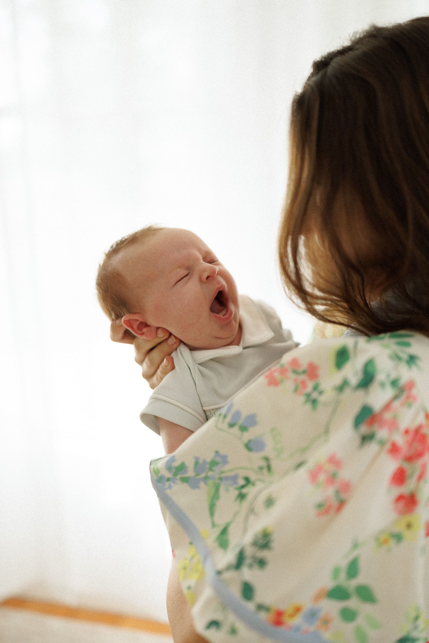 Mother holding yawning newborn baby in white onesie near bright window.