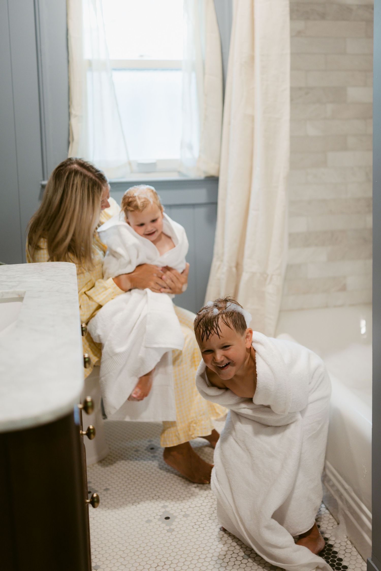 Family in white robes enjoying a cozy bathroom moment, with mother holding baby and father smiling nearby.