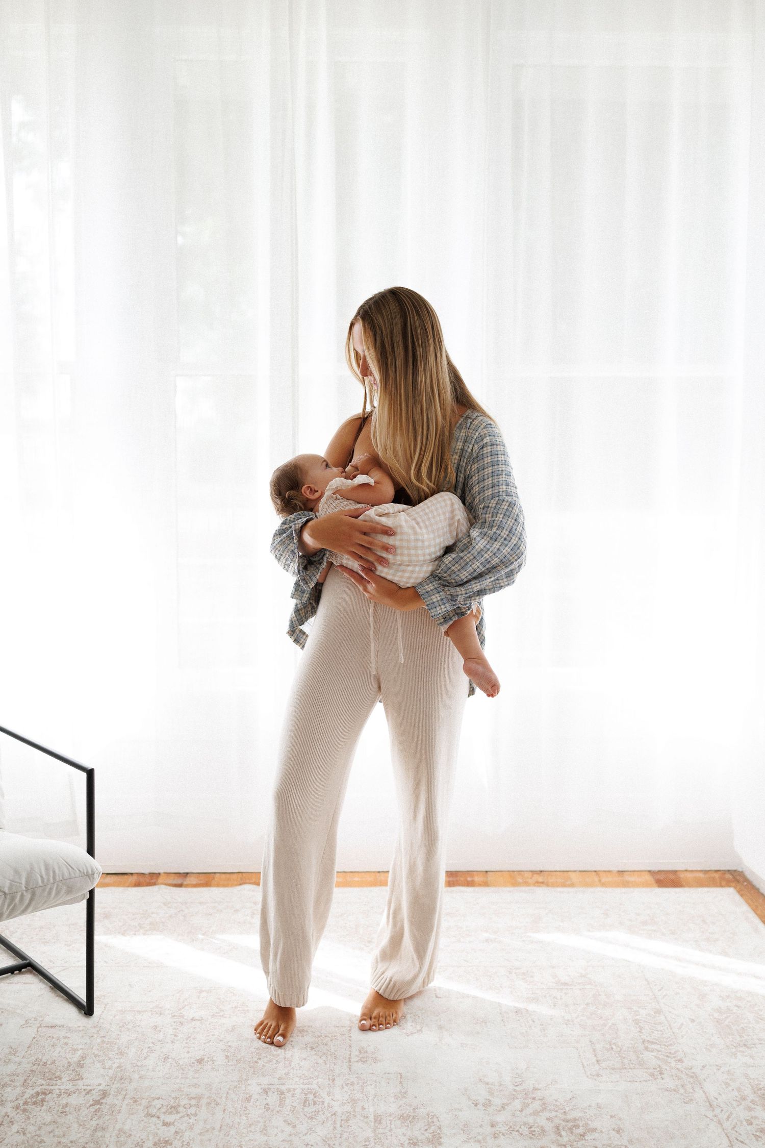 Person in white pants and grey sweater stretches on light carpet while practicing yoga movements in sunny room.