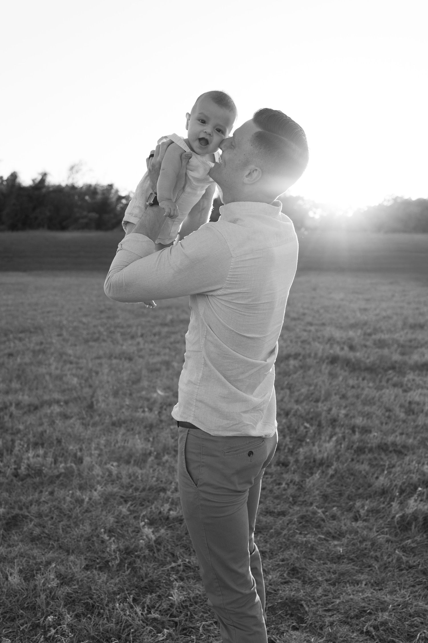 A tender black and white photo of a parent lifting a baby against a sunlit field during golden hour.