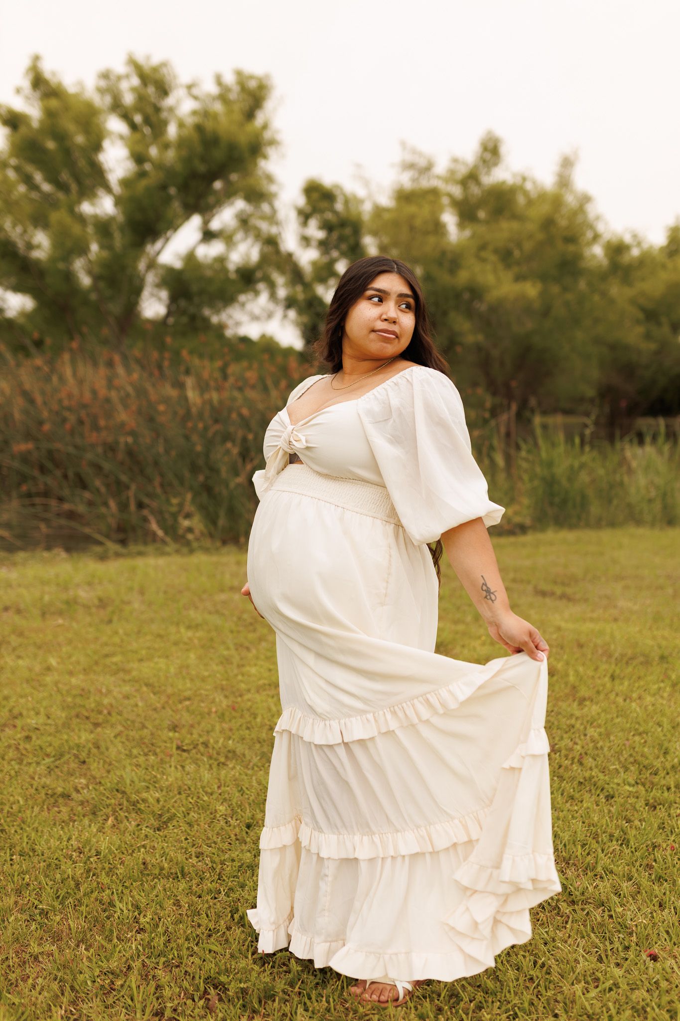 A person in a flowing white dress poses gracefully in a natural outdoor setting during a maternity photoshoot.