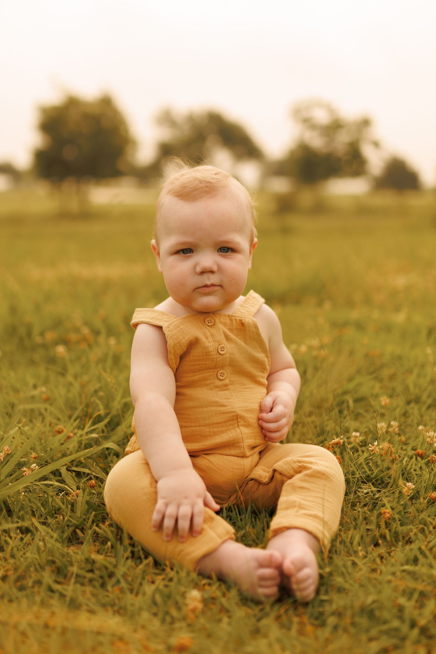 A baby in a yellow romper sits in a grassy field during golden hour.