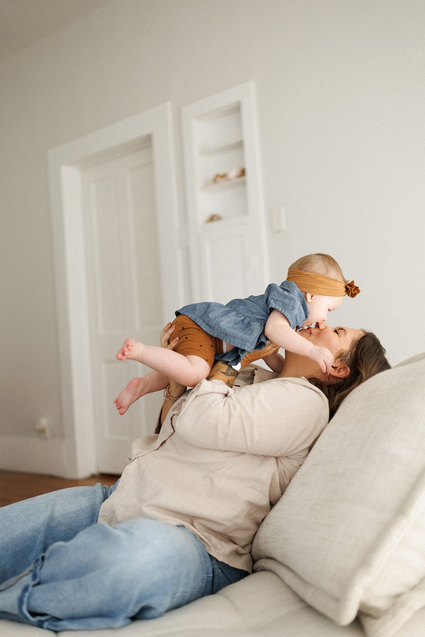Mother lying on sofa playfully lifting laughing toddler in denim dress above her.