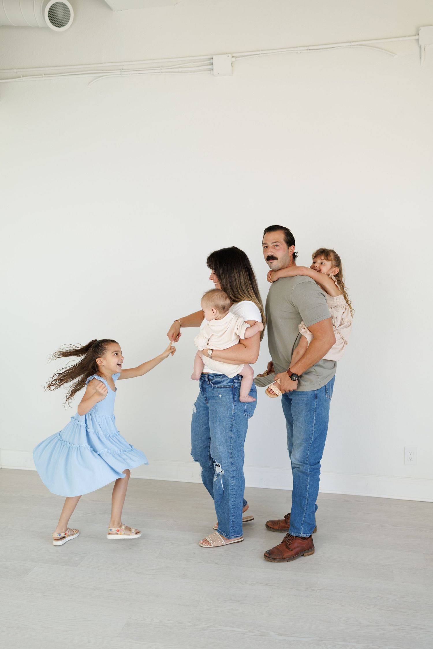 A family in casual attire plays together in a bright empty room, showing a sequence of playful interaction.