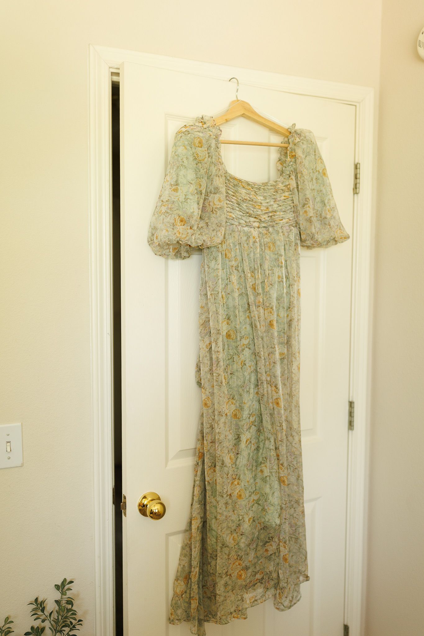 A vintage-style floral dress with puff sleeves hangs on a wooden hanger against a white door.