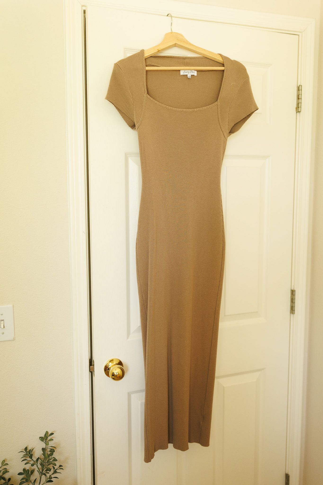 A tan ribbed bodycon maxi dress with short sleeves hangs on a white wooden hanger against a cream colored door.