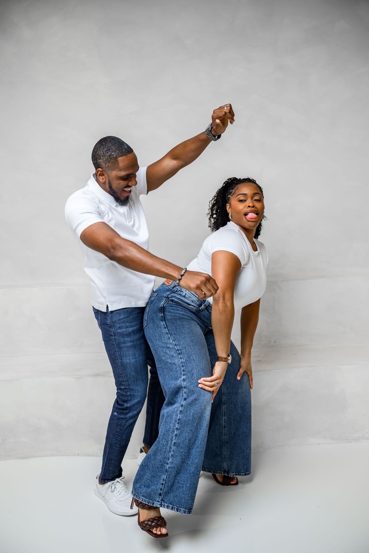A couple in matching white t-shirts and jeans dance together playfully against a gray background.