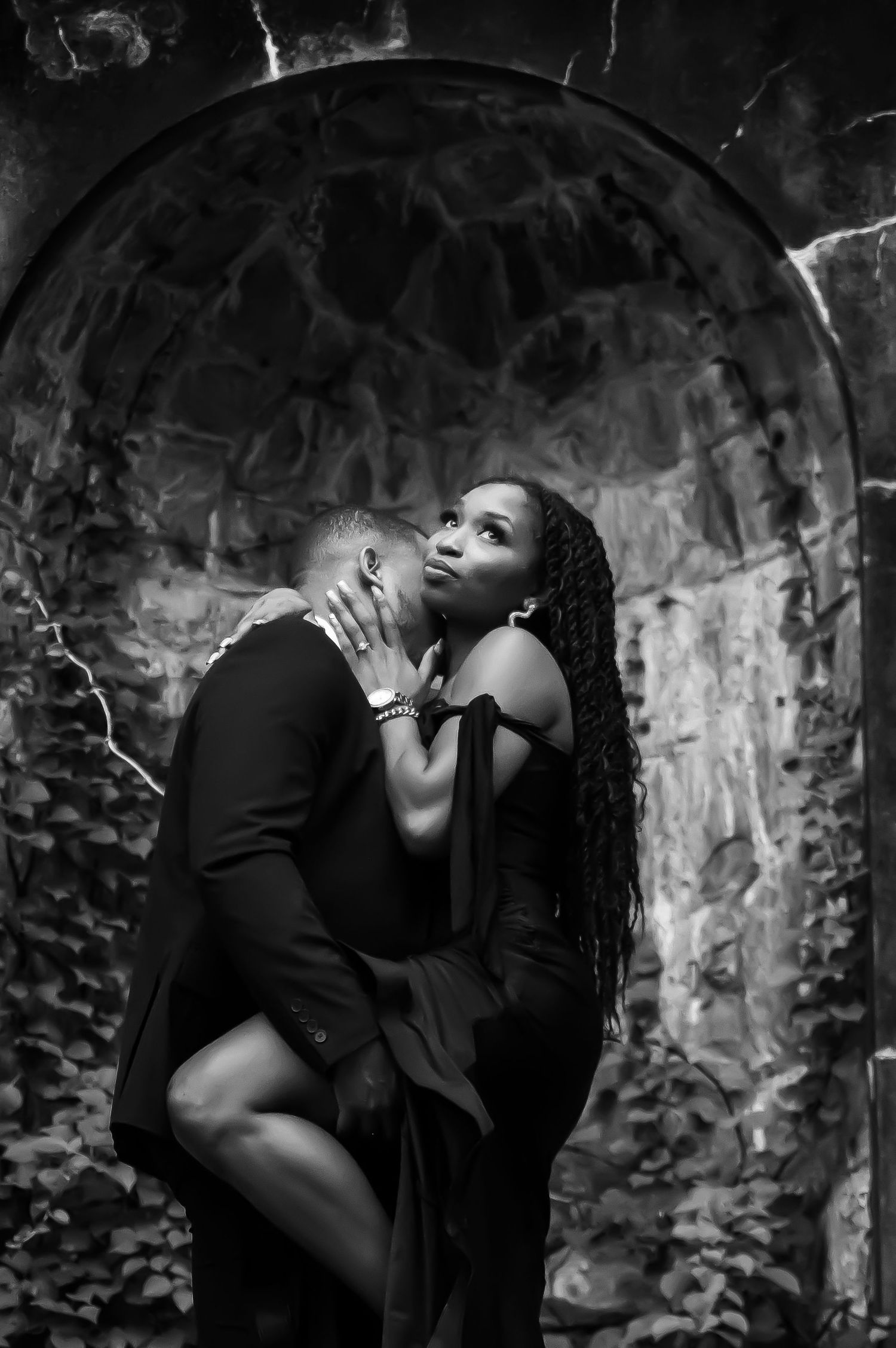 Romantic black and white portrait of couple embracing under stone archway.