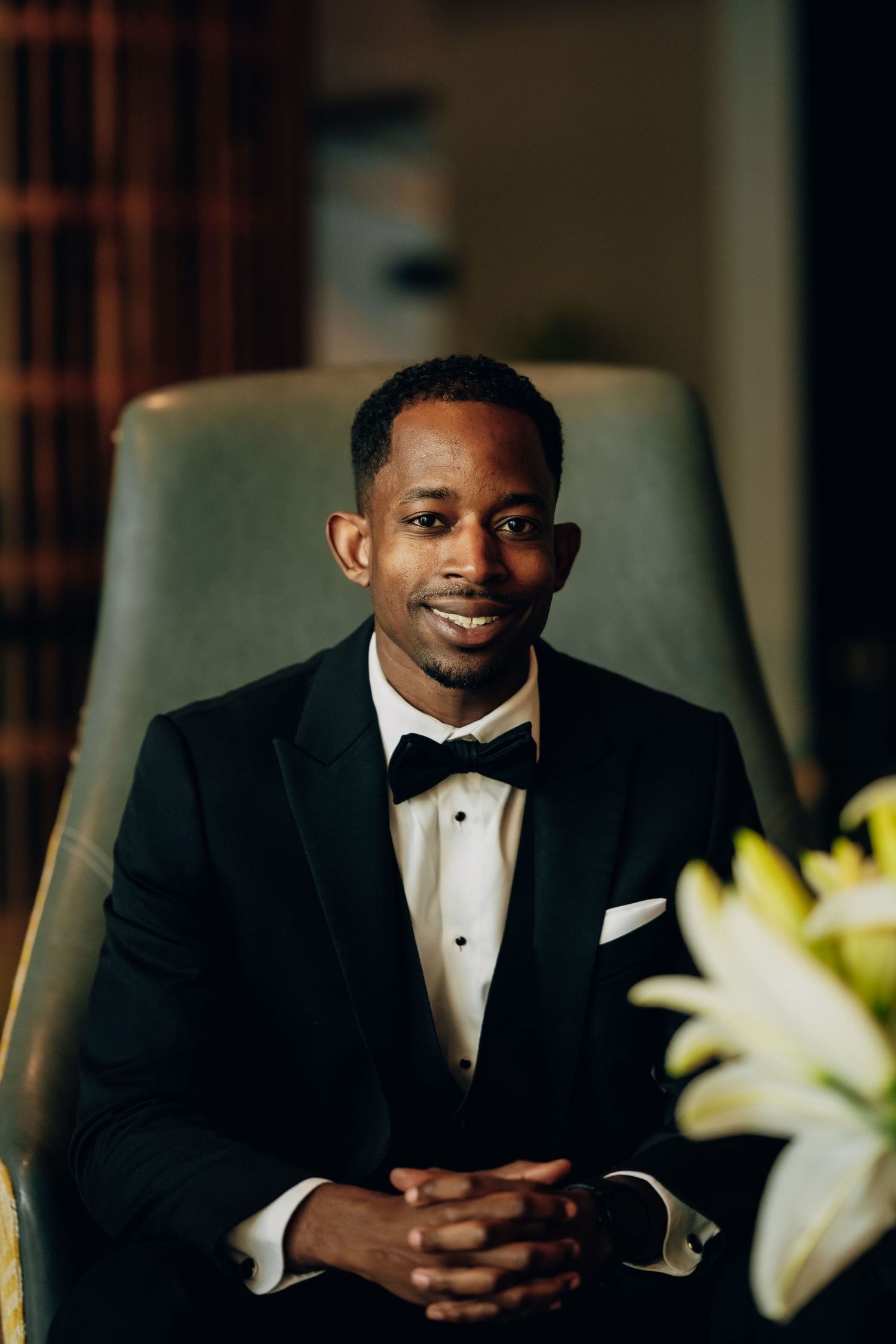 Portrait series of person in black tuxedo sitting with white lilies in background.