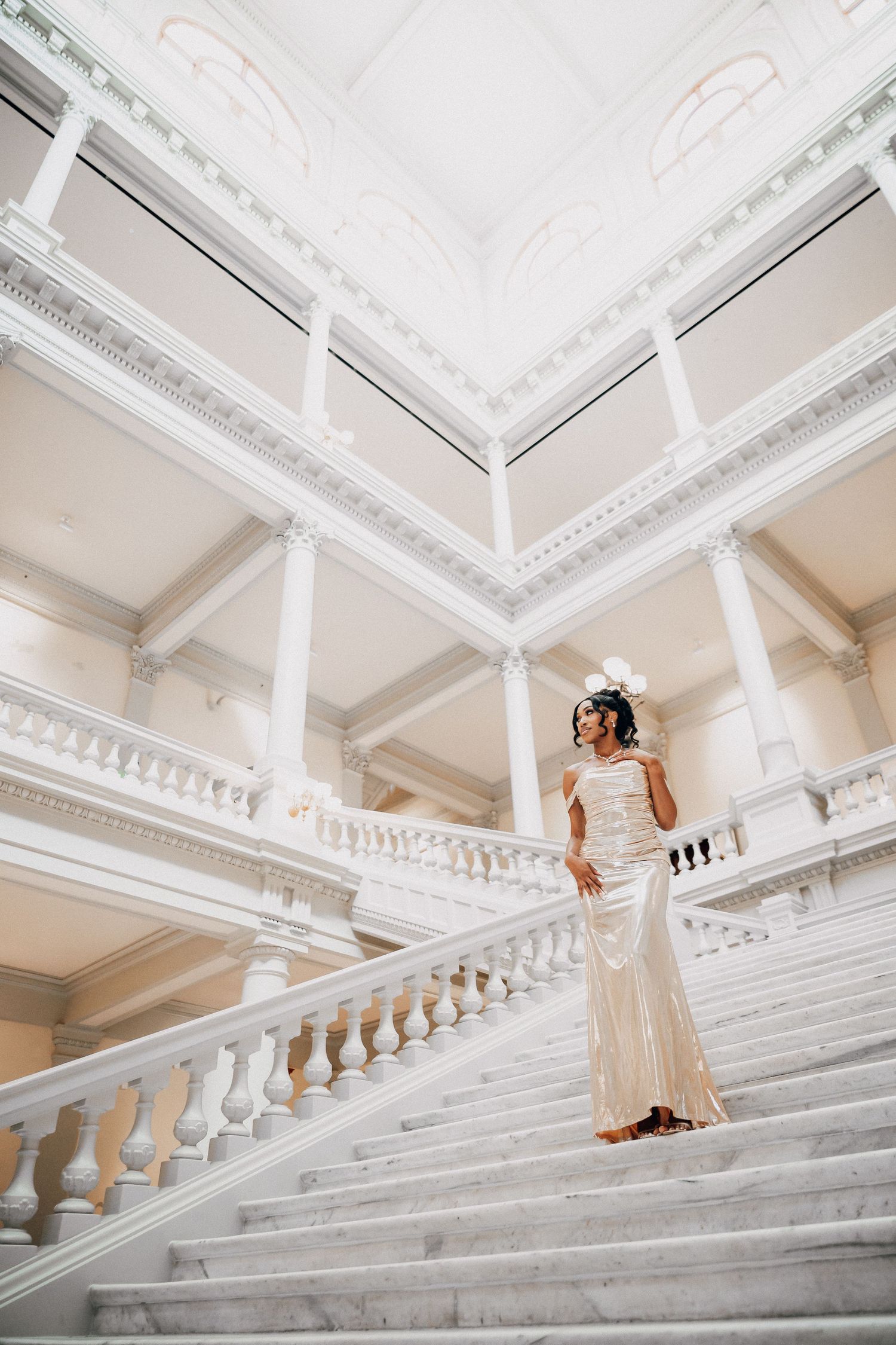 Person in white dress descending grand marble staircase with ornate white ceiling.