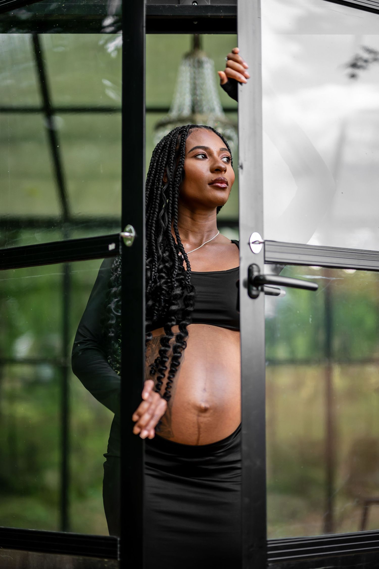 Person in black attire poses artistically in a glass greenhouse setting with natural lighting.