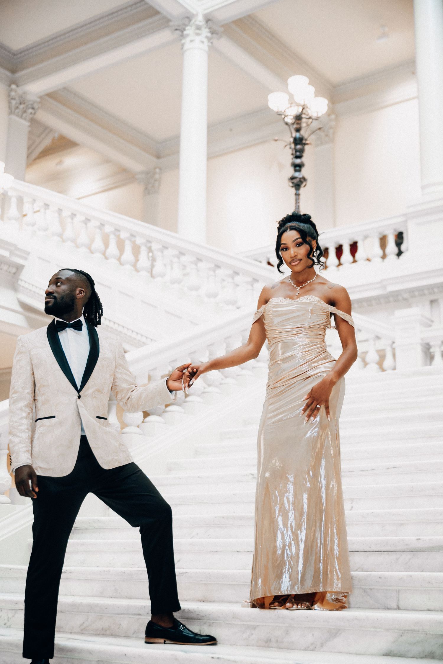 A couple poses elegantly on a grand white marble staircase, with the bride in a champagne gown and groom in white jacket.