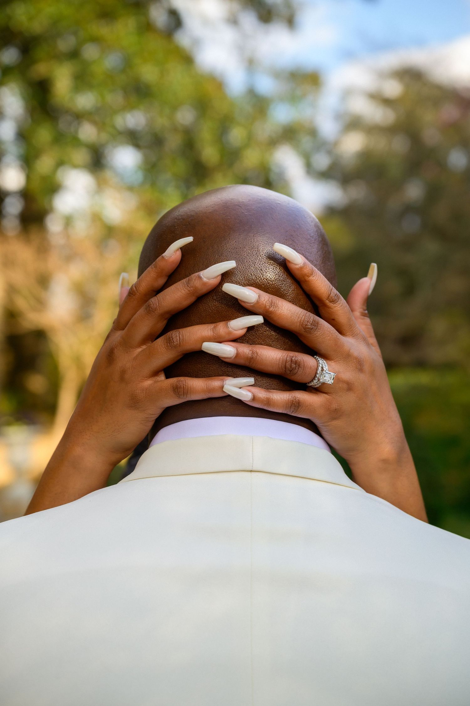 Woman's hands with long white manicured nails and wedding ring embrace the back of someone's head wearing white.