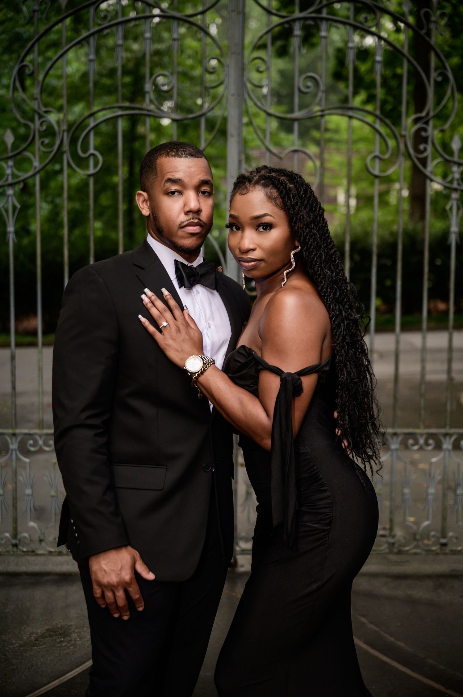 Elegant couple in black formal wear pose playfully by decorative metal gates.