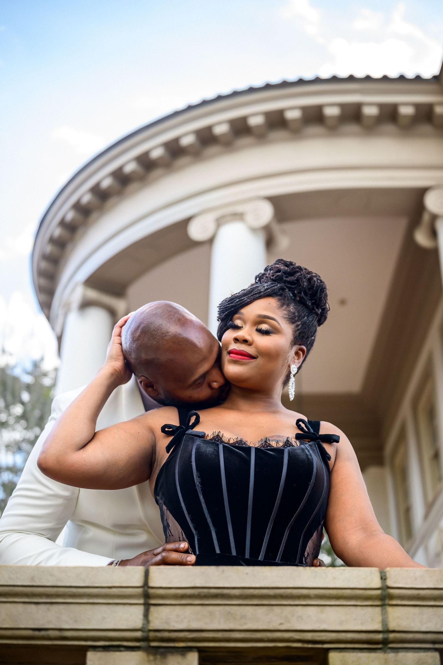A couple in elegant attire share intimate moments on an ornate balcony during a romantic photoshoot.