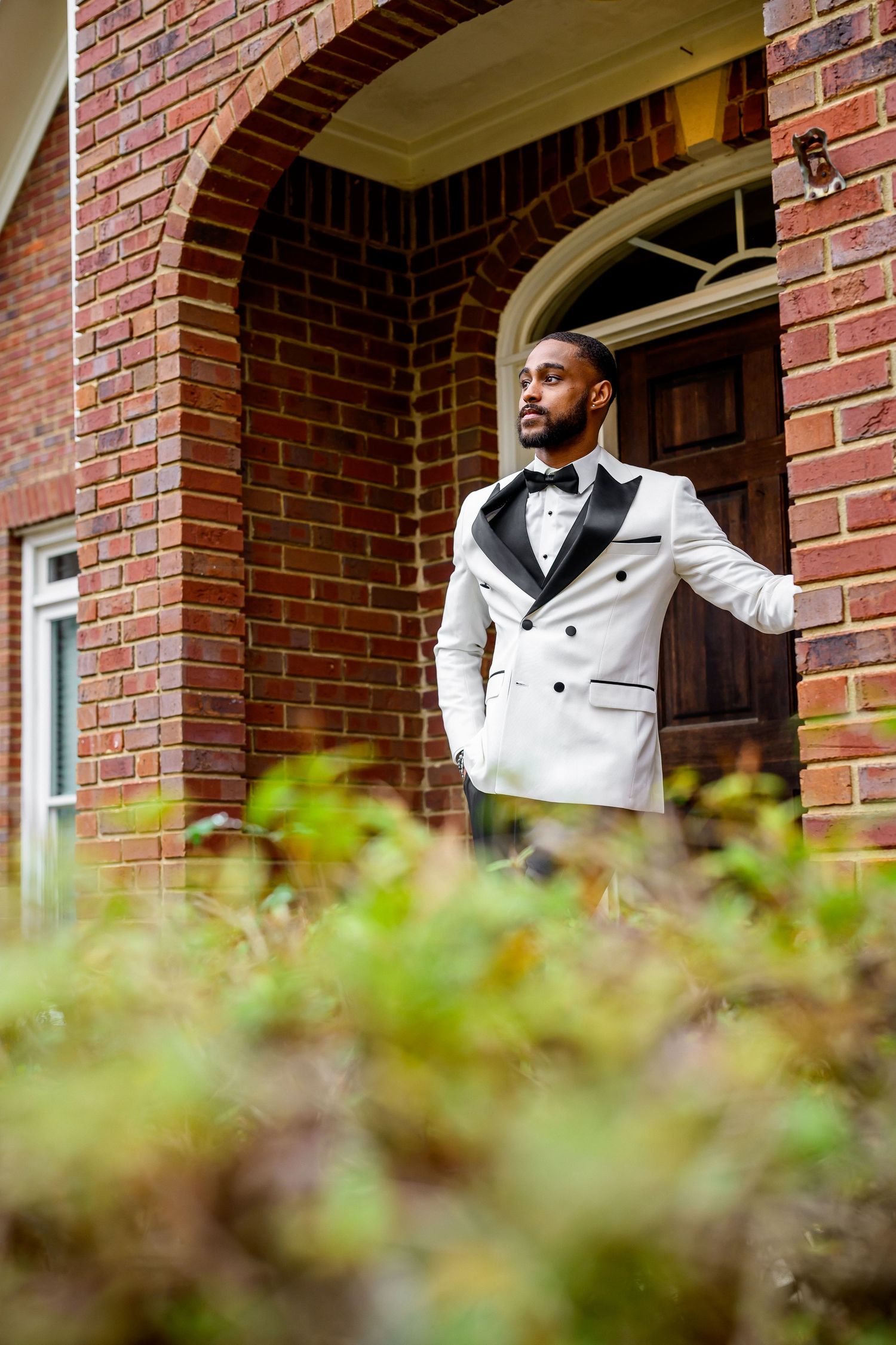 A person in a white tuxedo jacket and bow tie poses elegantly against a brick building entrance with greenery in foreground.