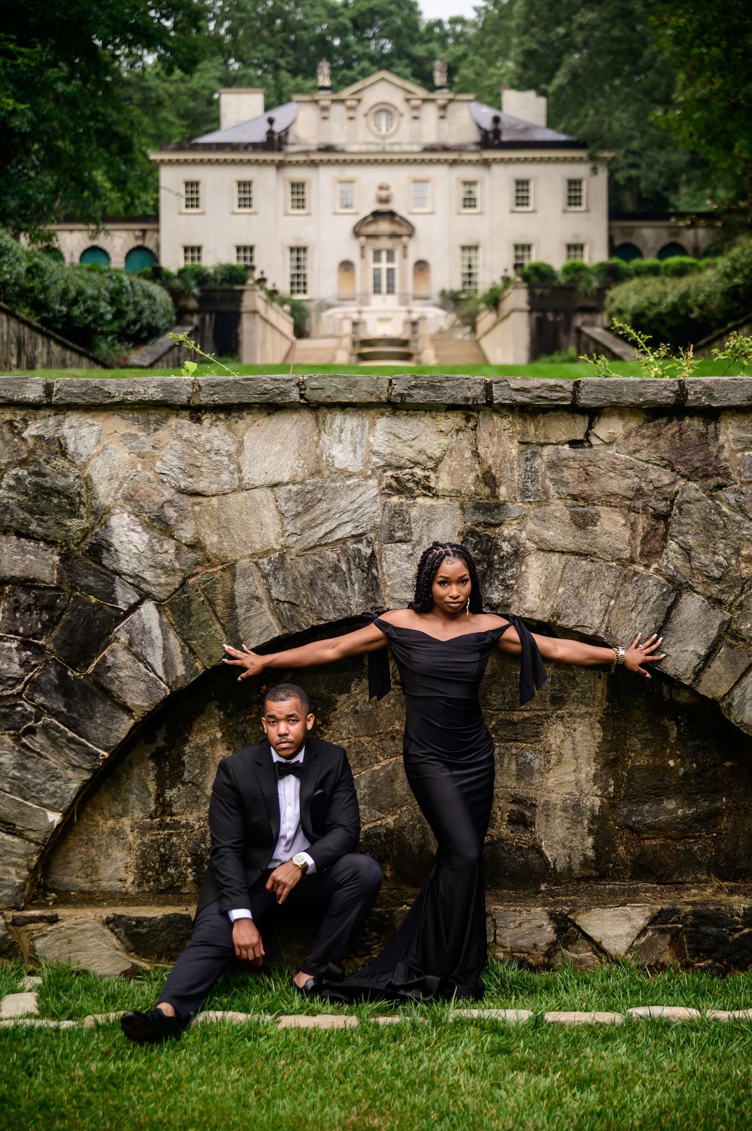 Stylish couple strikes artistic poses by historic stone architecture and manicured lawn.