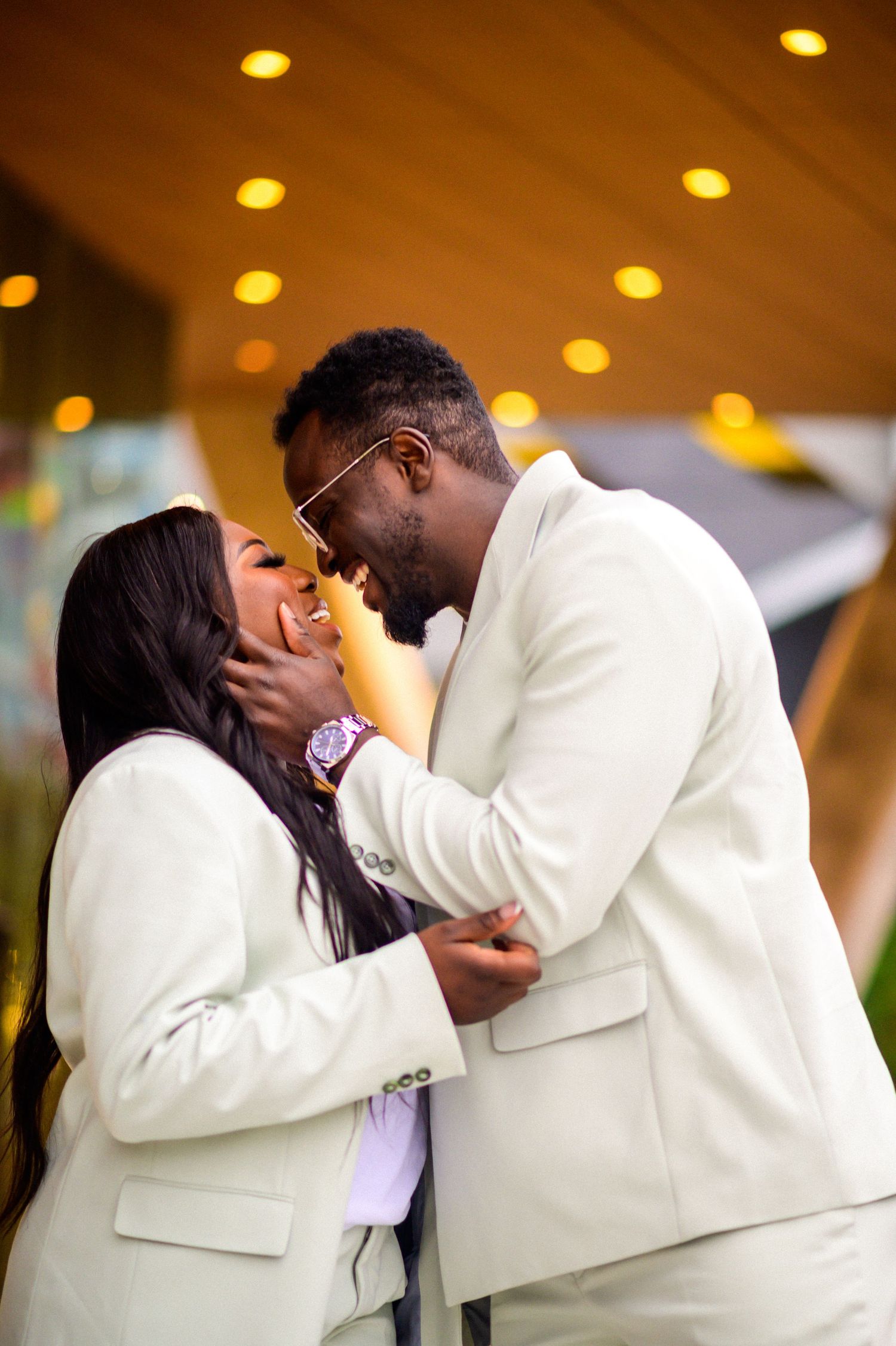 Couple shares tender moments in white formal wear under warm indoor lighting.