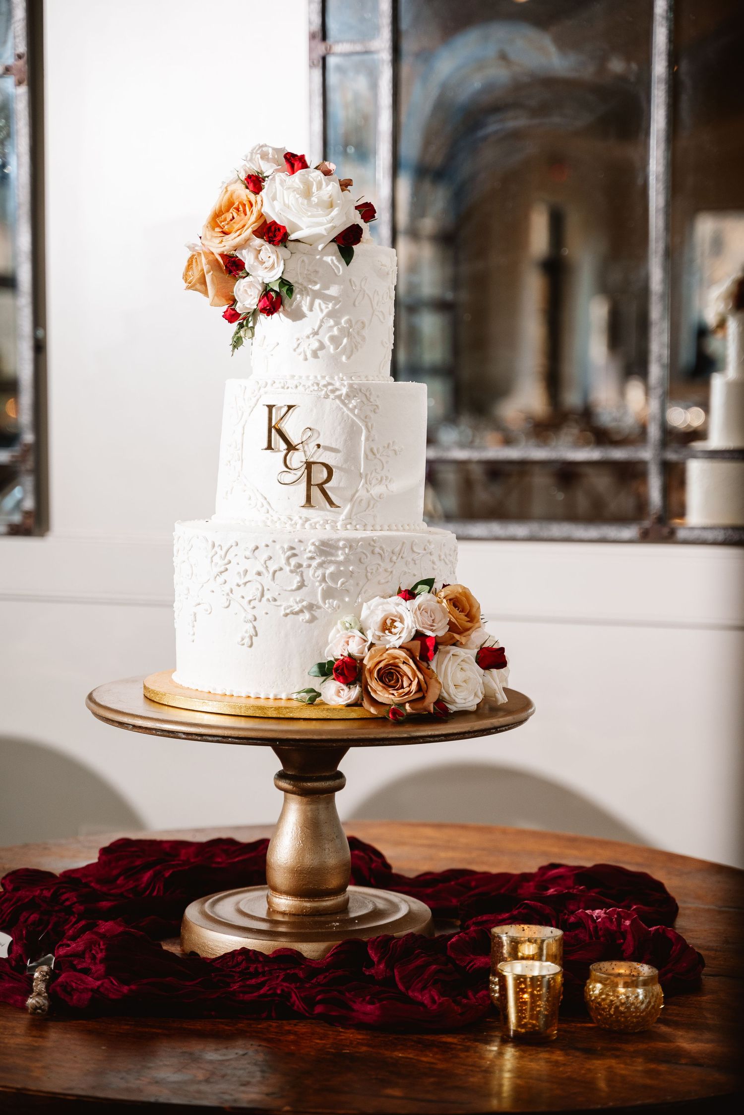 White wedding cake decorated with peach and burgundy roses on gold cake stand with velvet accents.