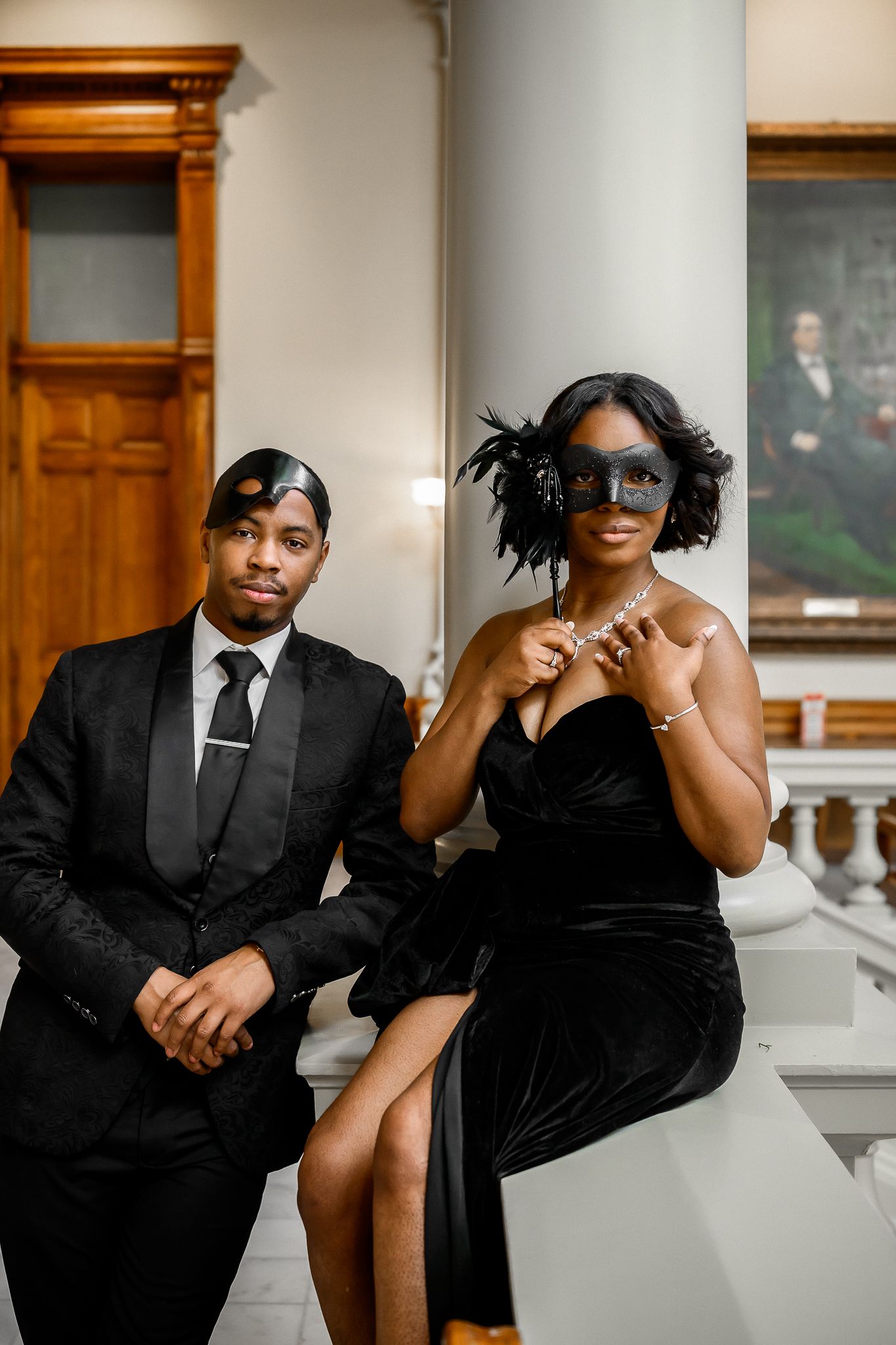 Elegant couple in formal black attire pose together in a grand historic building interior.