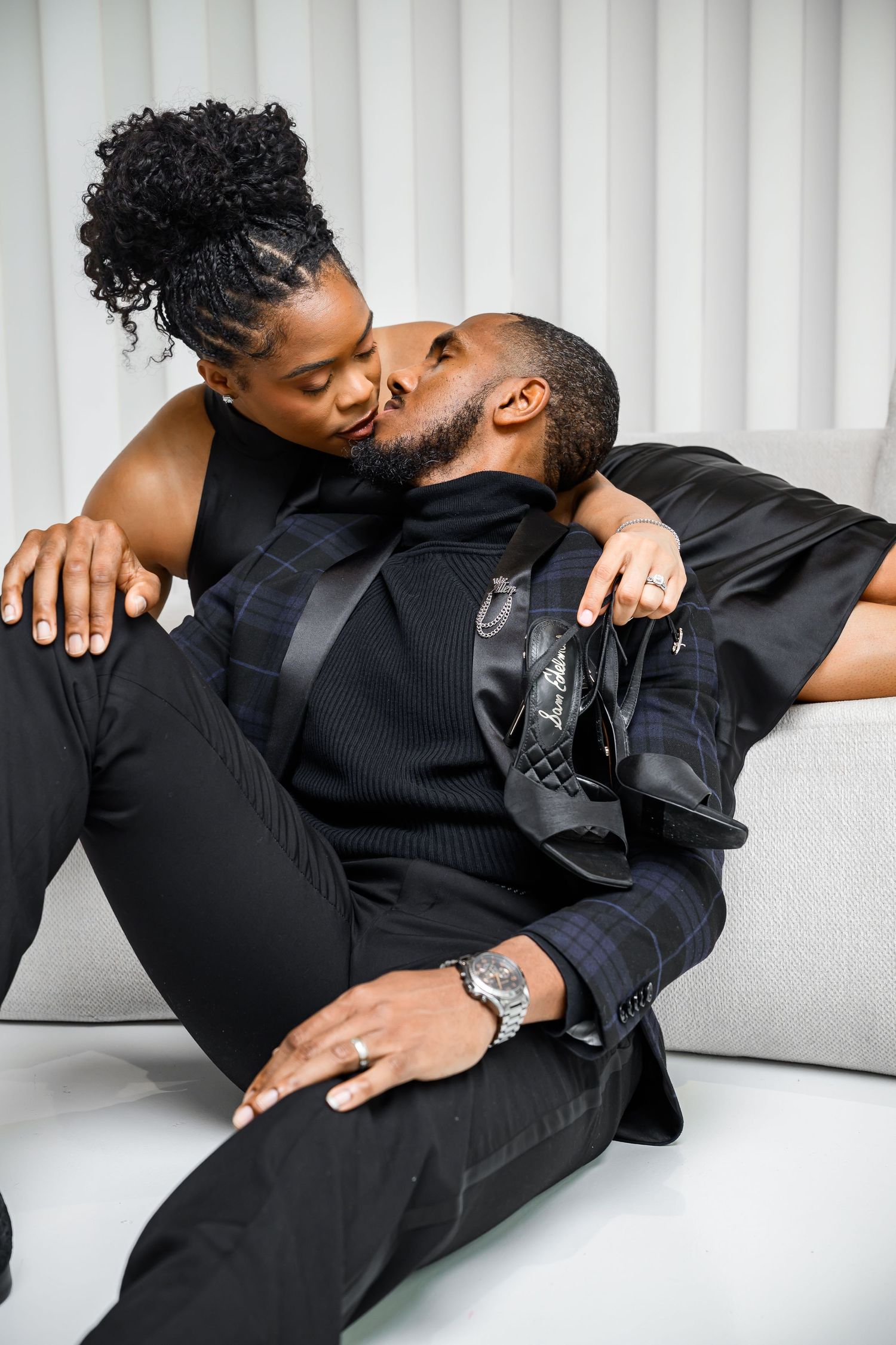 A couple in black outfits sharing an intimate moment while lounging on a white couch.