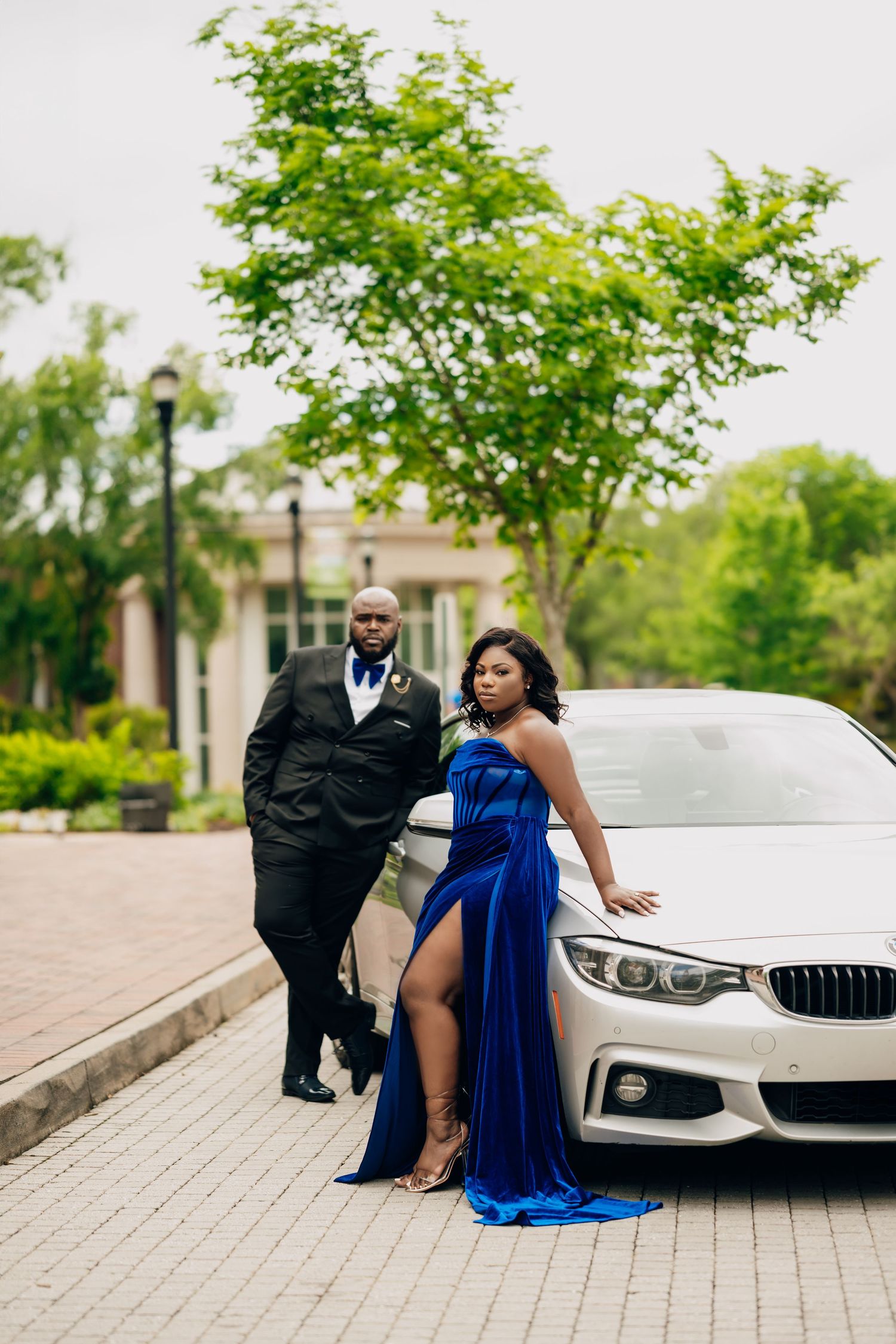 Elegant couple in formal wear poses next to a white luxury BMW for engagement photos.