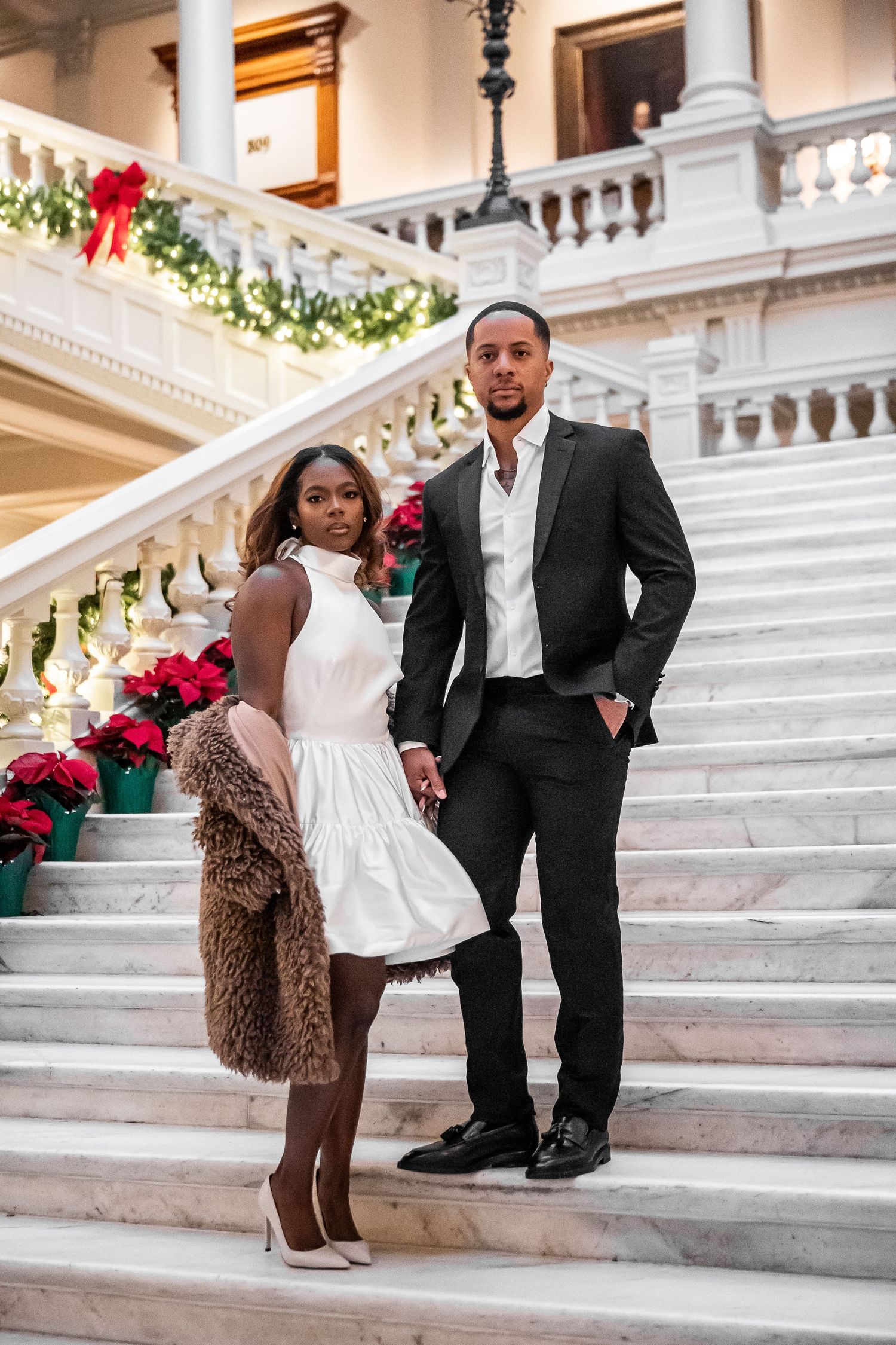 Elegant couple poses on grand marble stairs decorated with red poinsettias.