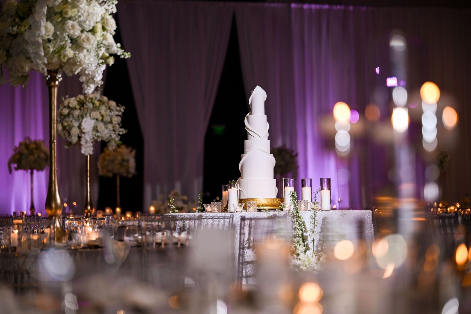 Elegant white wedding cake illuminated by candlelight with purple-lit draping in background.