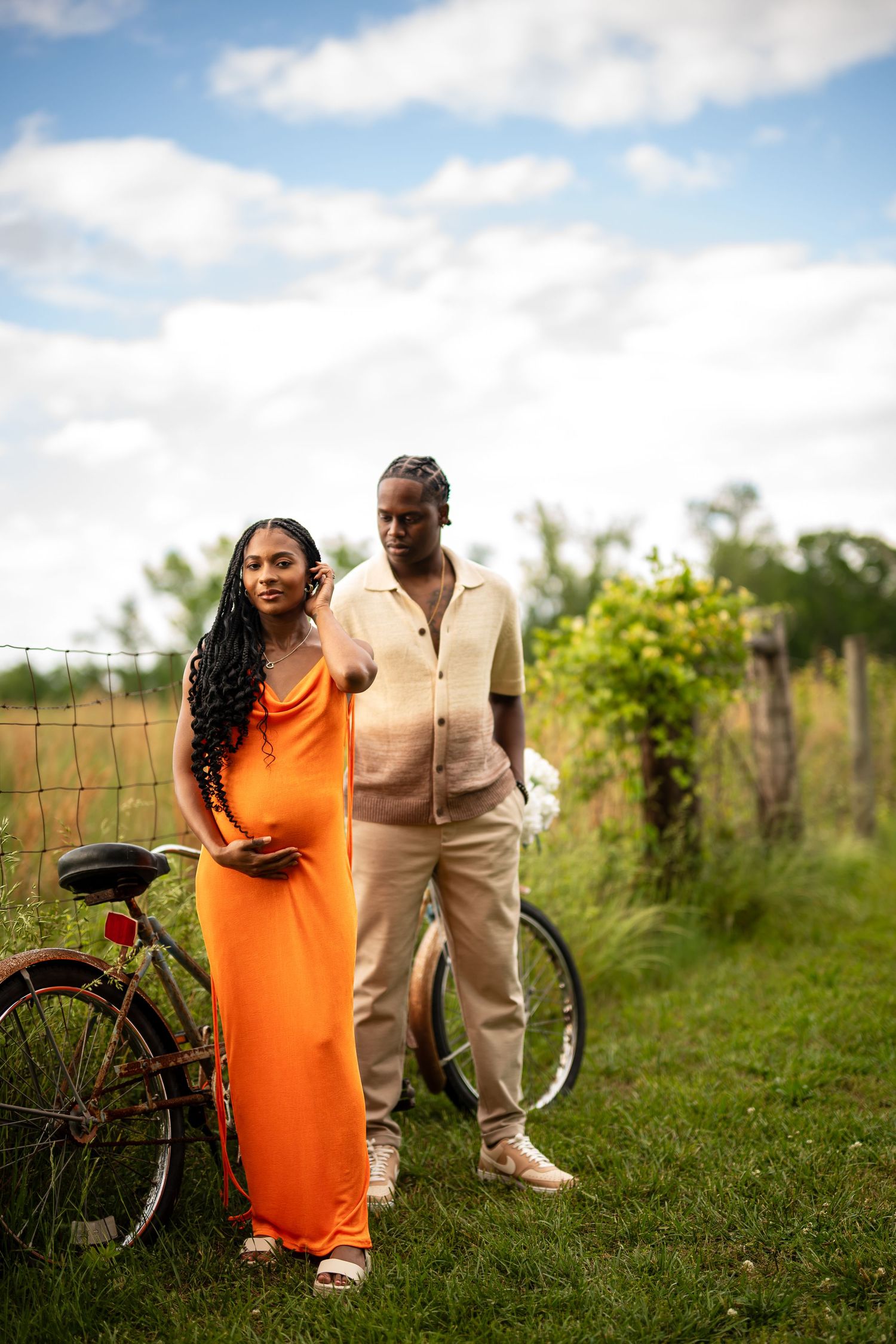 Couple in orange dress and beige outfit pose together in a sunny garden with vintage bicycle.