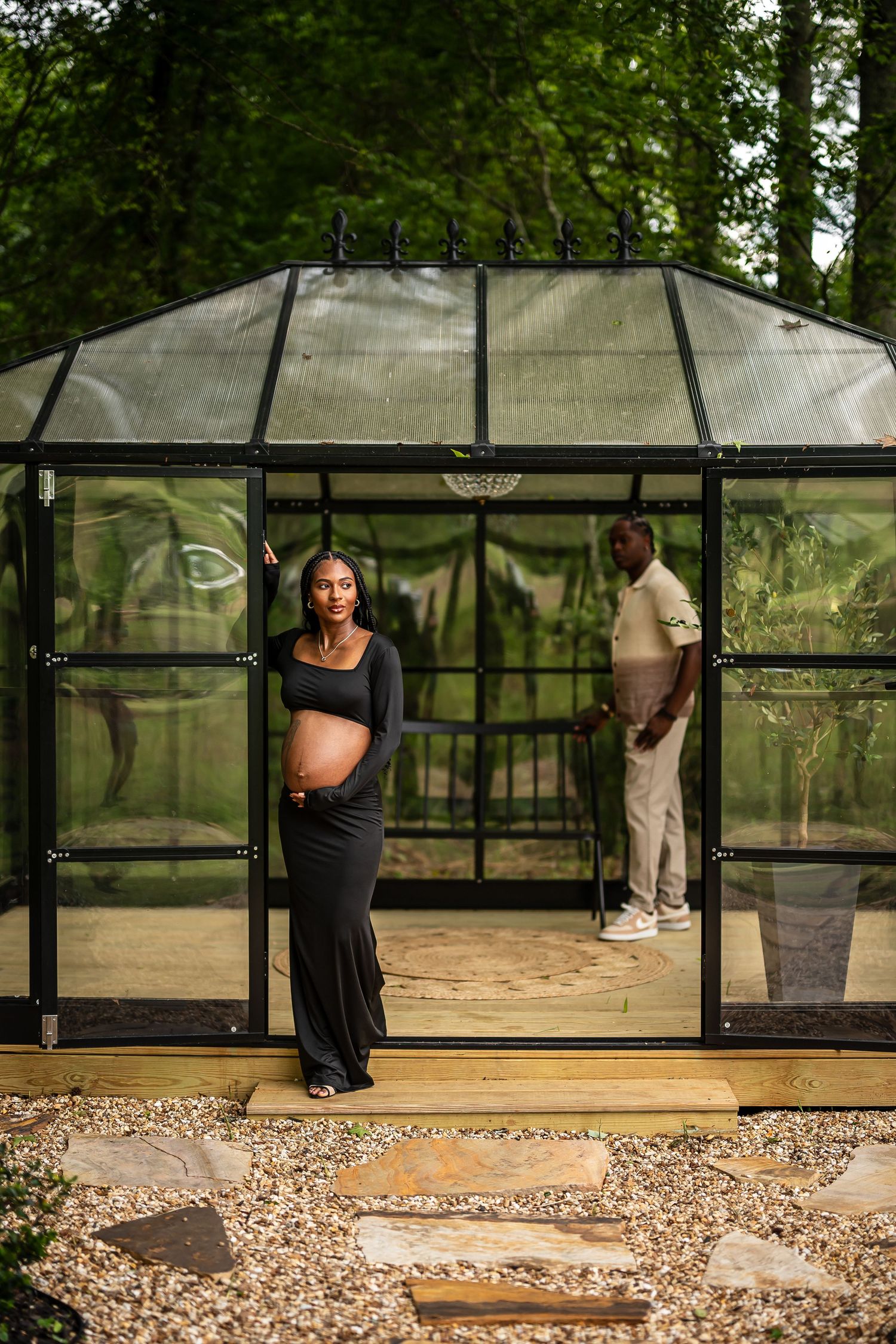 A glass greenhouse with black frame sits on a gravel path surrounded by lush greenery.