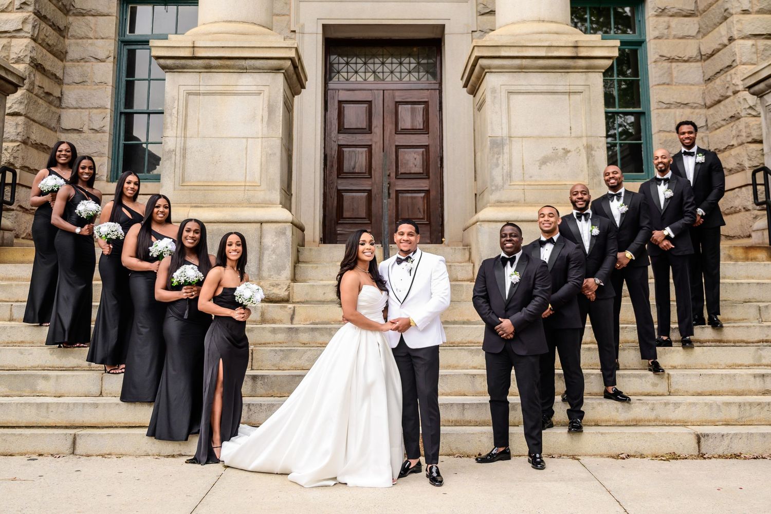 Wedding party poses on stone steps of historic building in formal black and white attire.